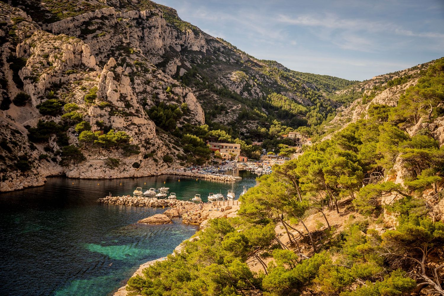 Scenic coastal cove with small boat harbor nestled between rocky cliffs and green pine trees along Mediterranean coastline.