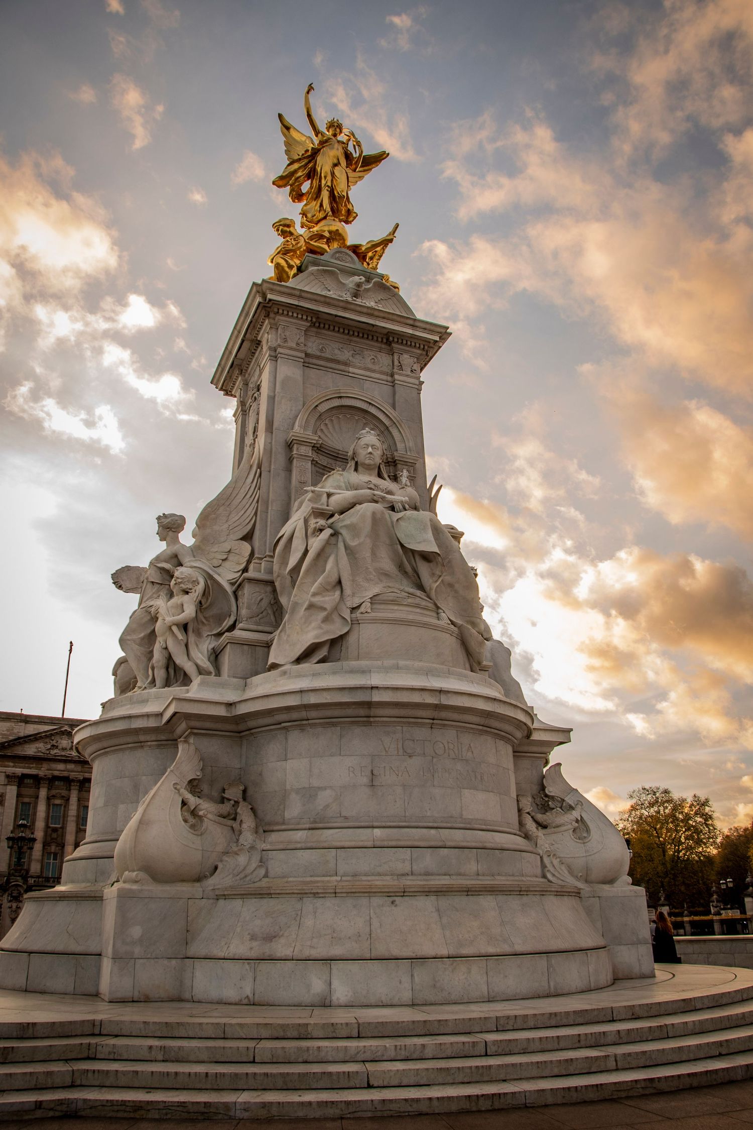The Victoria Memorial monument in London features marble statues and gold-topped figures against a dramatic sunset sky.