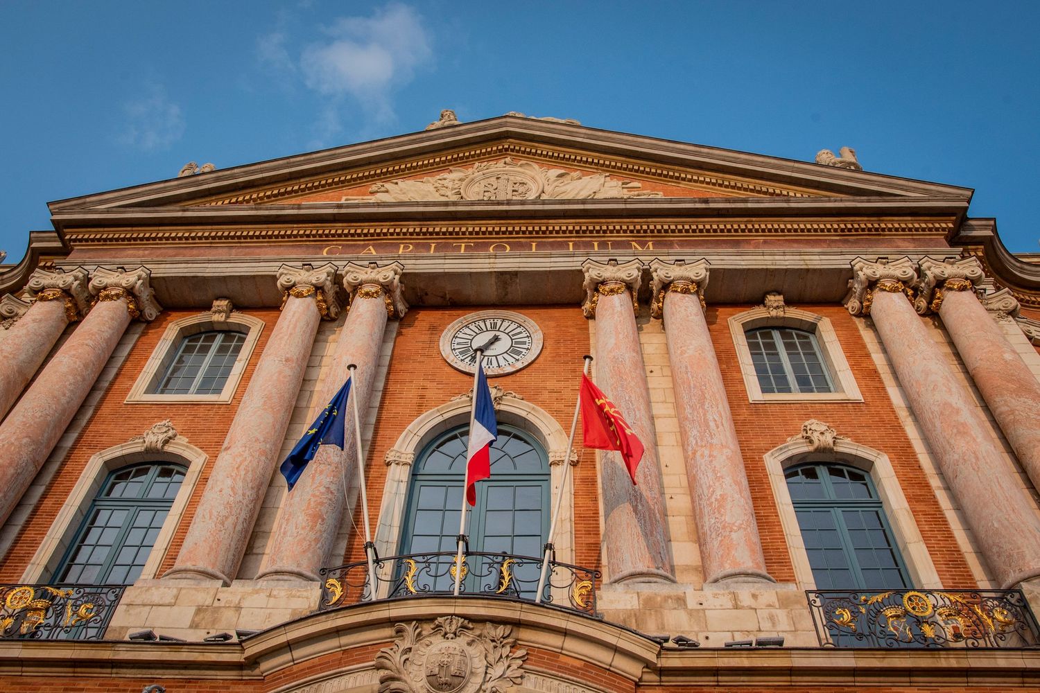Historic red-brick building facade with triangular pediment and classical columns displaying French and European flags in Toulouse.