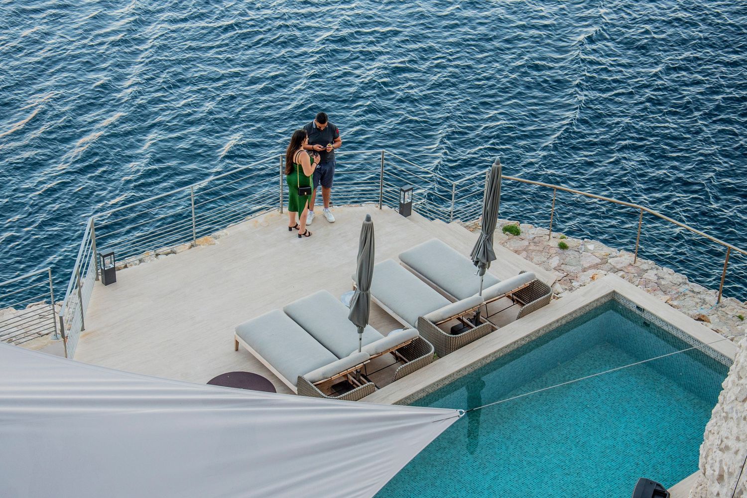 Aerial view of a luxurious infinity pool overlooking blue ocean waters with lounge chairs on a white sundeck.
