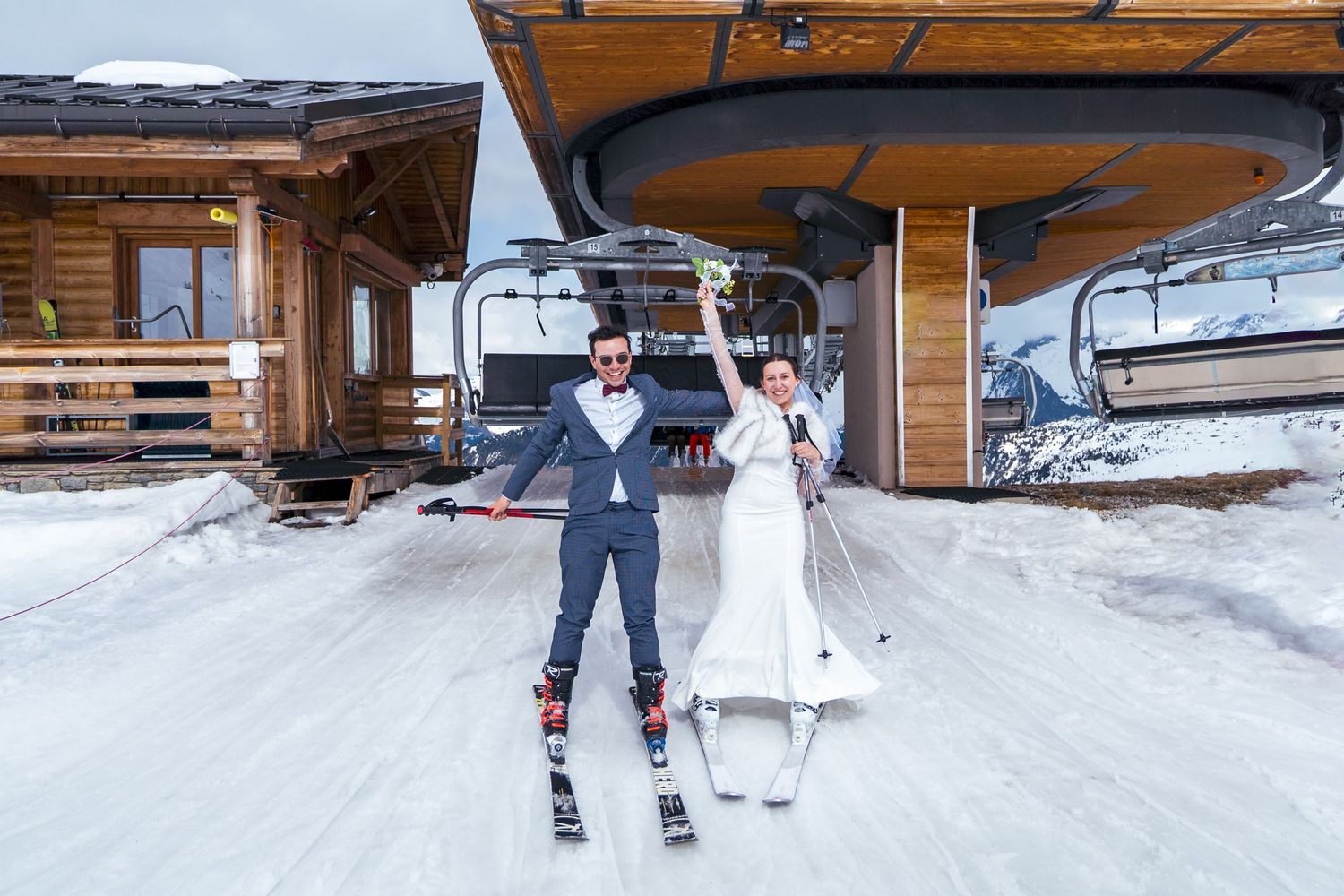 Two skiers holding hands while riding a ski lift at a snowy mountain resort with wooden chalets visible.