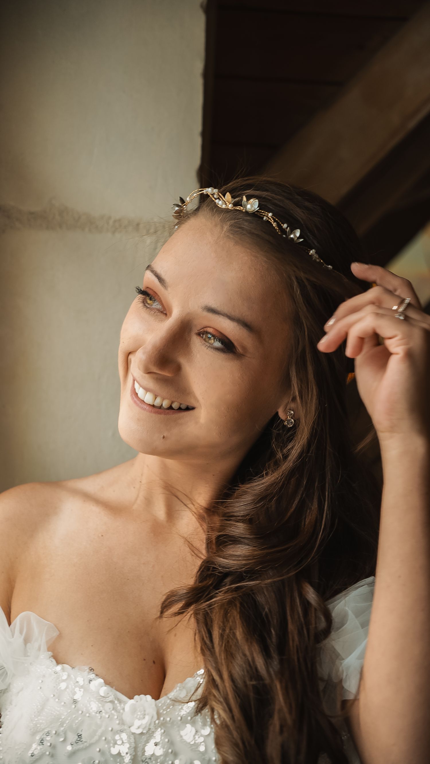 A bride in a white strapless wedding gown wearing a delicate floral headpiece smiles warmly in natural lighting.