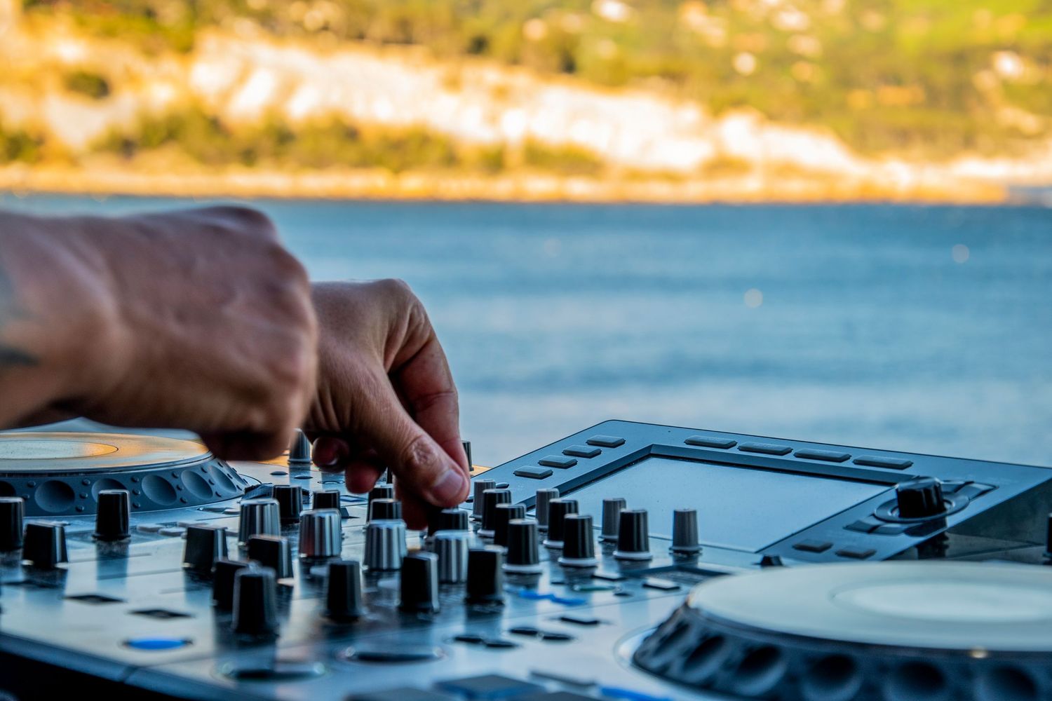 Close-up view of hands adjusting knobs on a DJ mixer controller against a blurred lakeside background.