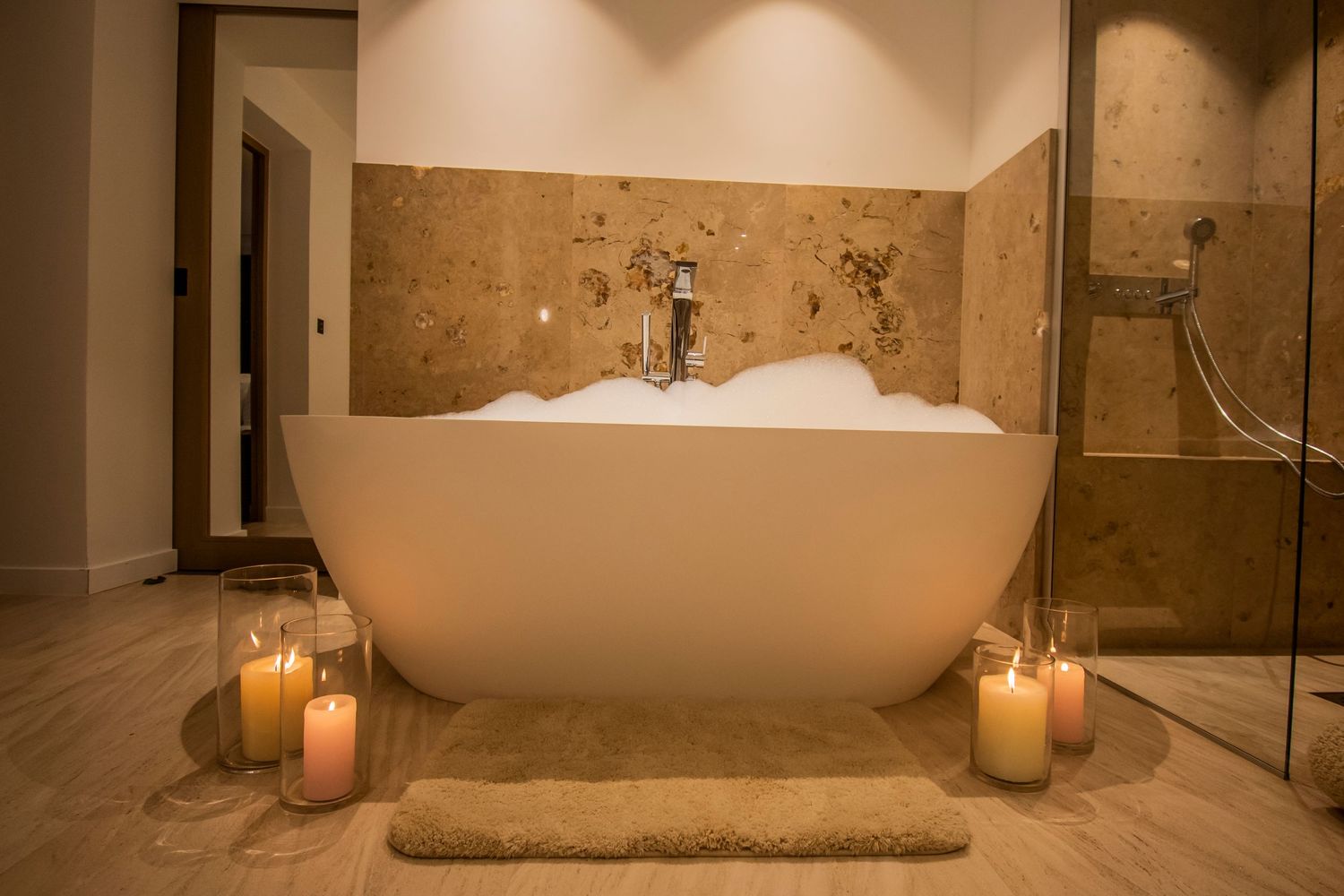 Modern freestanding white bathtub surrounded by glowing candles on beige marble floor in luxurious spa-like bathroom.