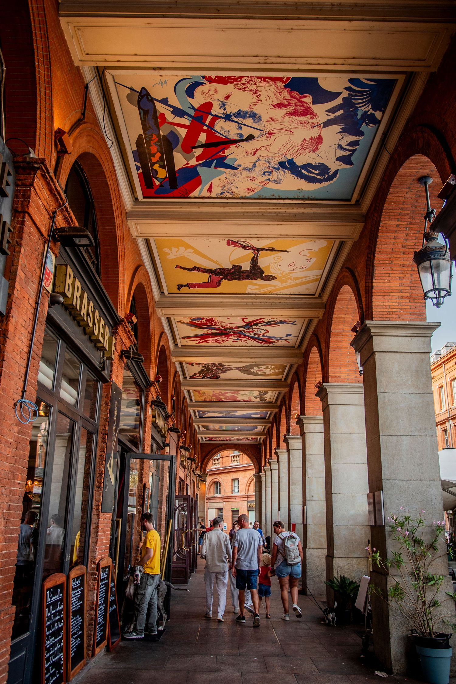 Historic arcade walkway with painted ceiling murals and red brick columns in Plaza Mayor, Madrid showing people strolling beneath.
