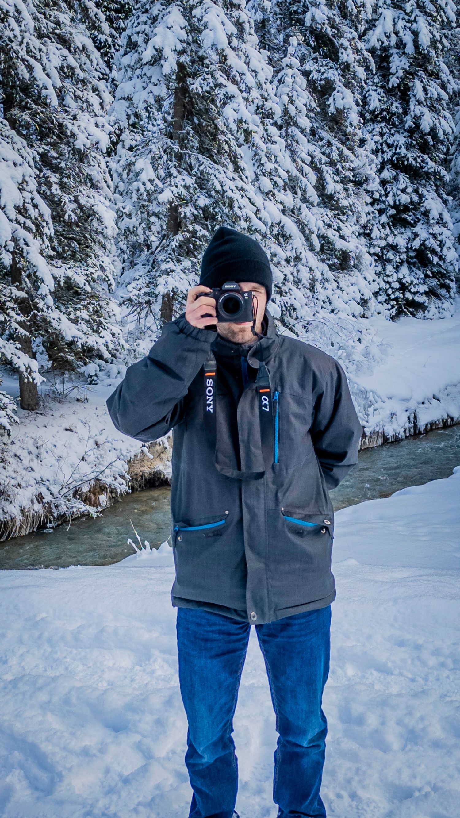 A photographer in winter gear takes pictures beside a snowy creek with frost-covered evergreen trees in the background.