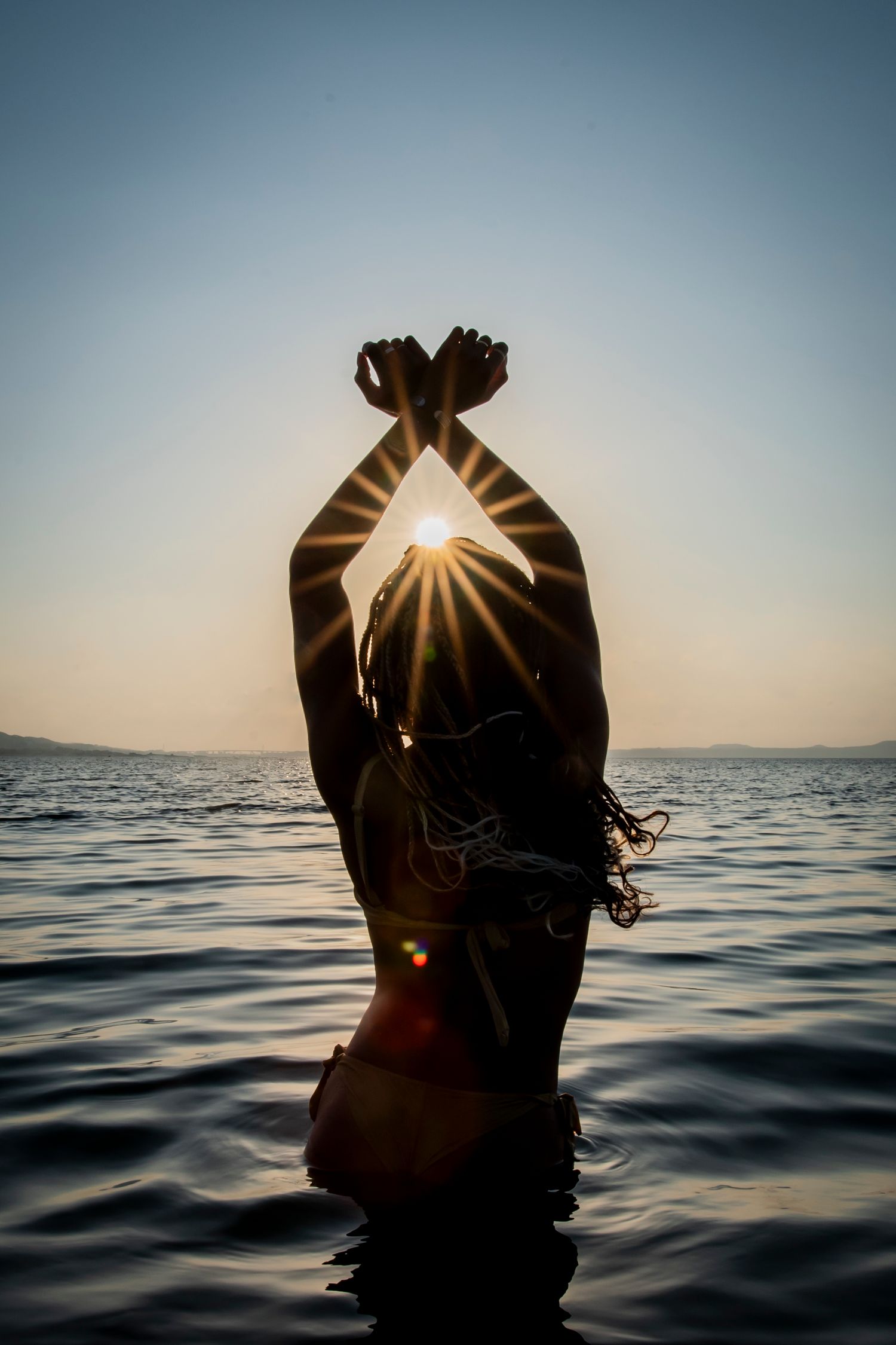 A silhouetted figure in water raises their hands overhead framing the setting sun through their fingers at sunset.