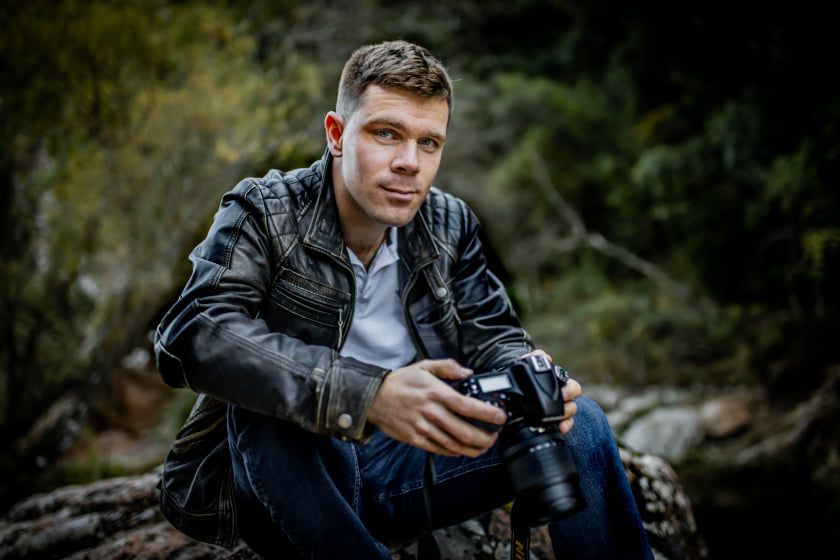 Figure in dark winter attire holding camera equipment while capturing shots in moody woodland setting.