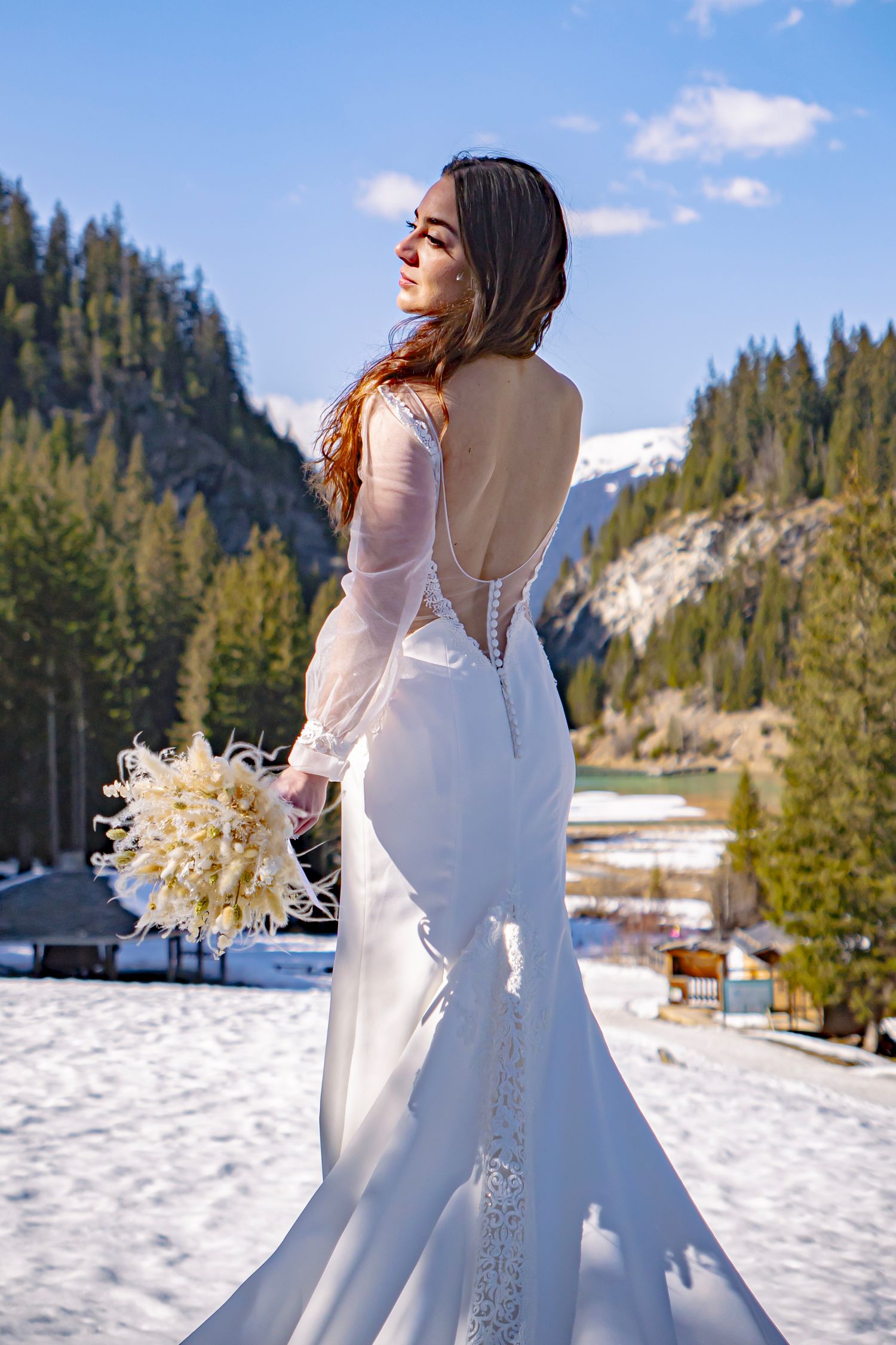 Bride in backless white wedding dress holds dried flower bouquet against snowy mountain landscape with evergreen trees.