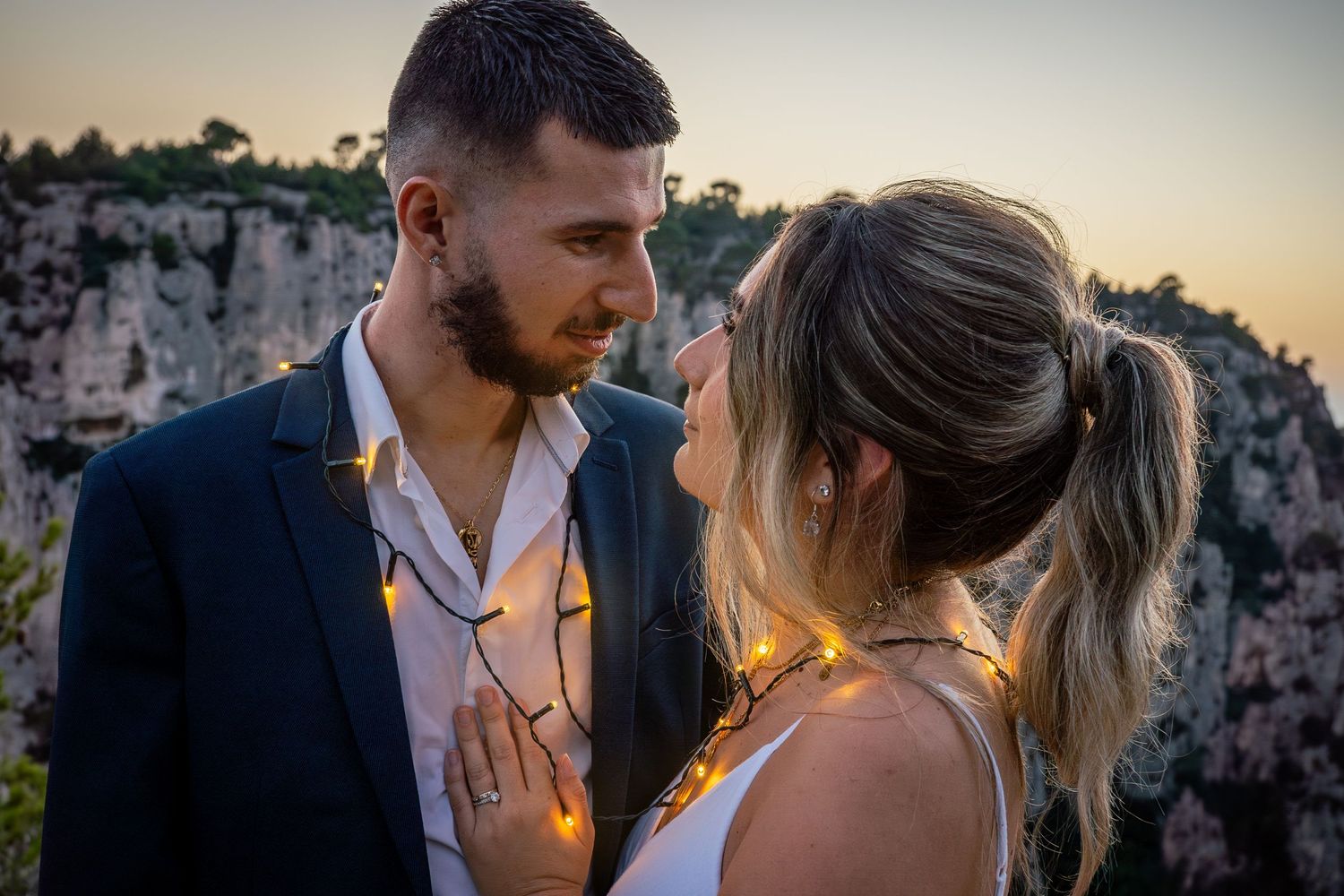 A romantic couple embraces during golden hour against a scenic rocky cliff backdrop at sunset.