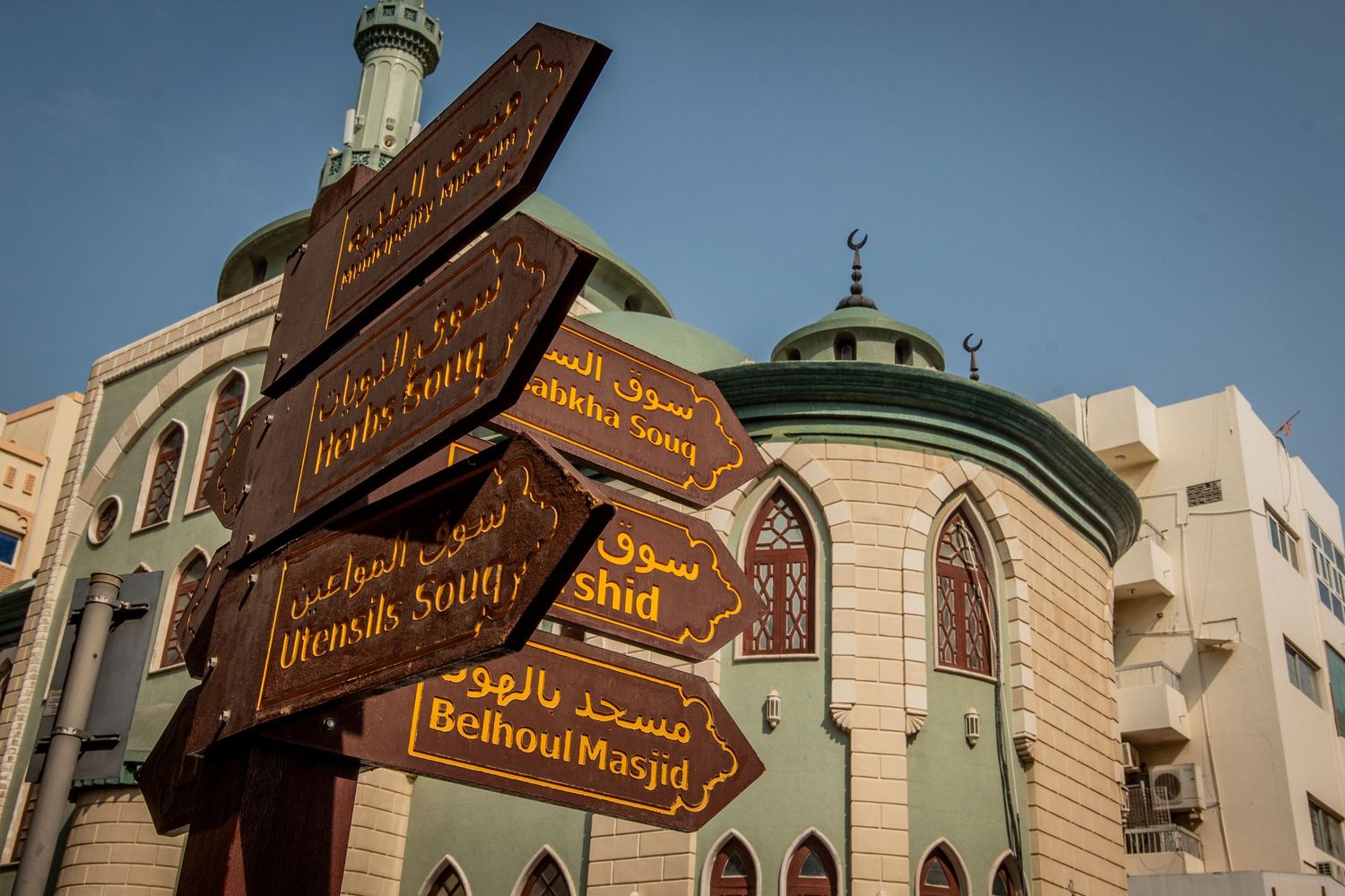 Wooden directional signs point to various locations outside a mosque with a green dome and traditional Islamic architecture.