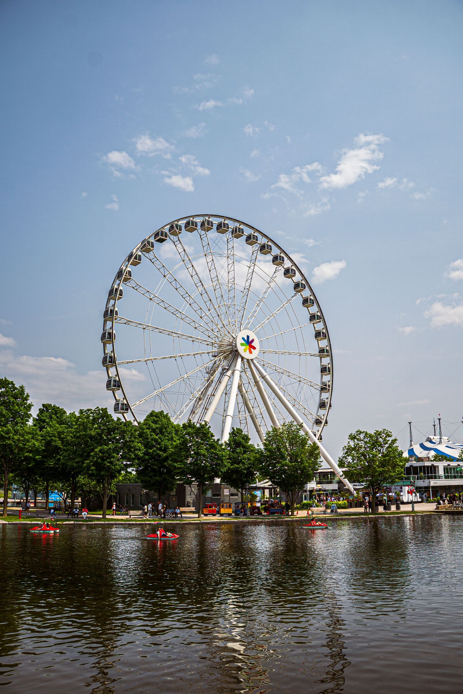 Large Ferris wheel rises above a lake with paddle boats in the foreground at an amusement park on a sunny summer day.