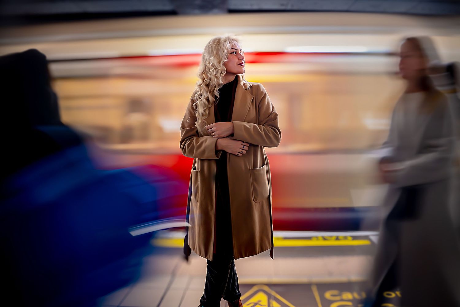 Woman in brown coat standing in subway station with motion blur in background.