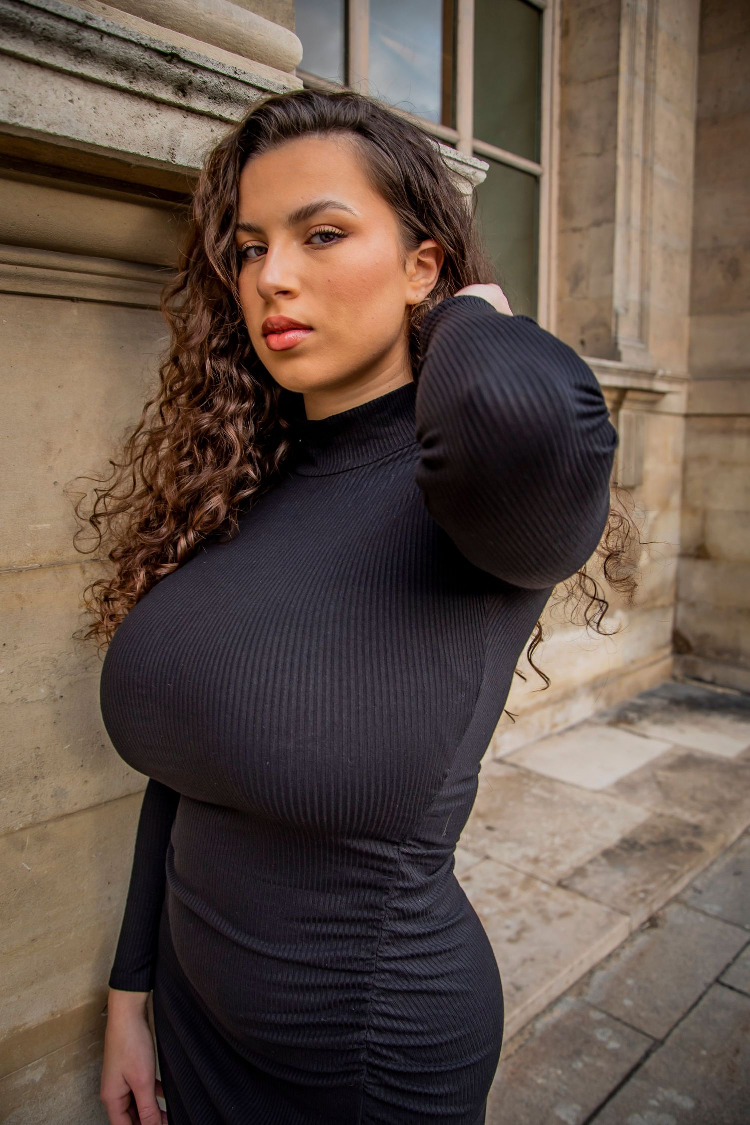 Model in black ribbed turtleneck dress poses near stone building with long curly hair and golden hour lighting.