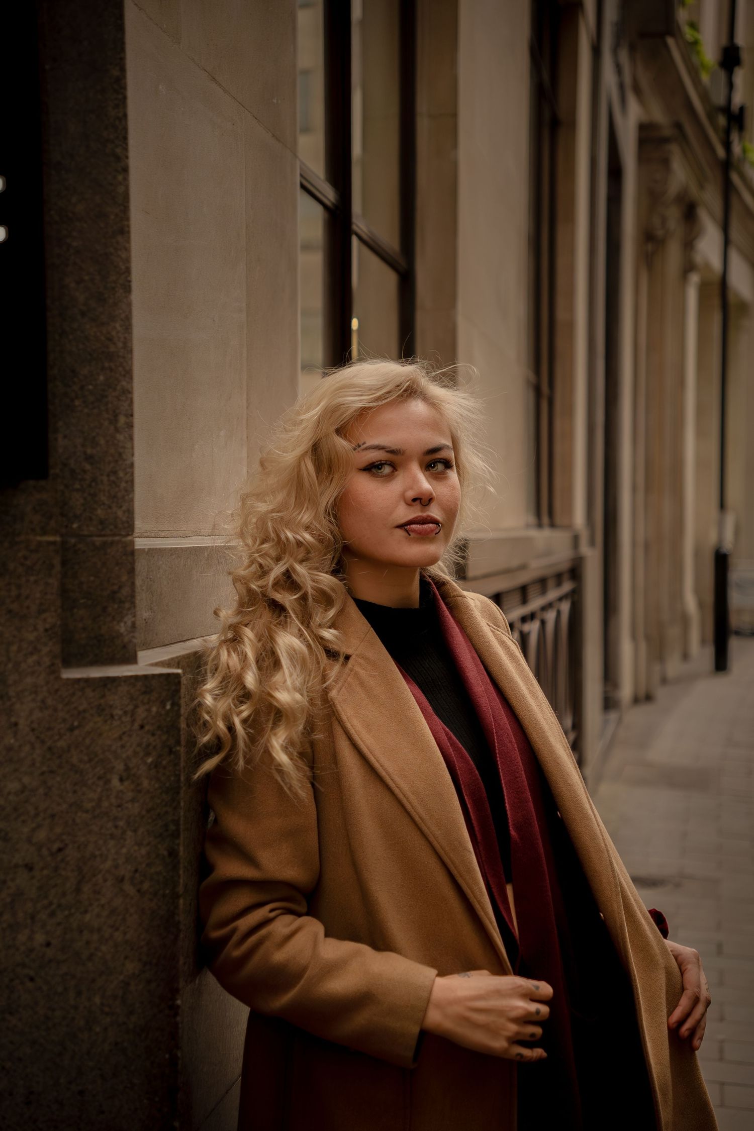 Stylish blonde in camel coat and black sweater poses against urban building exterior on cloudy day.