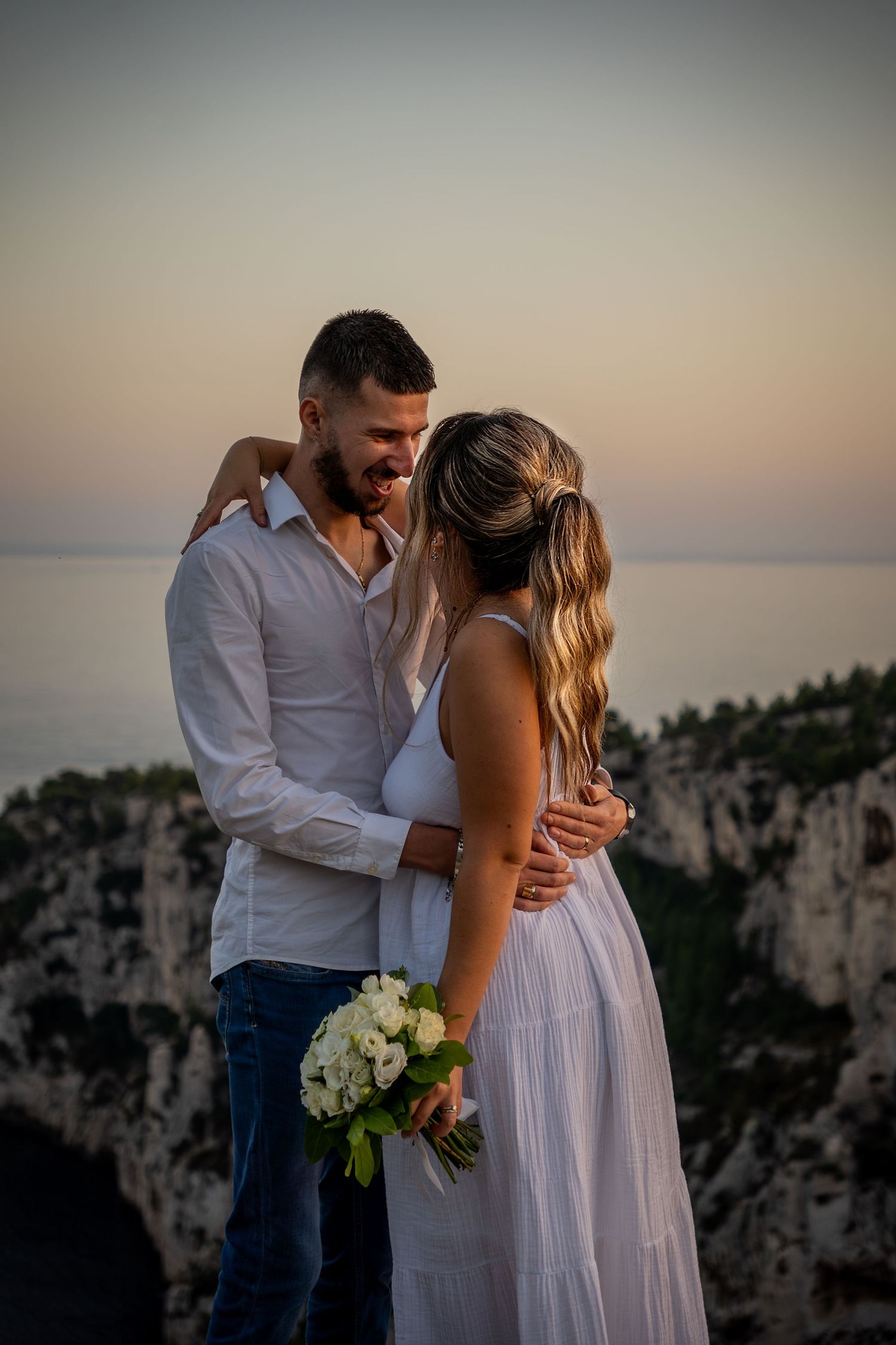 Romantic couple embraces at sunset on mountain cliff overlooking scenic ocean vista with white flower bouquet.