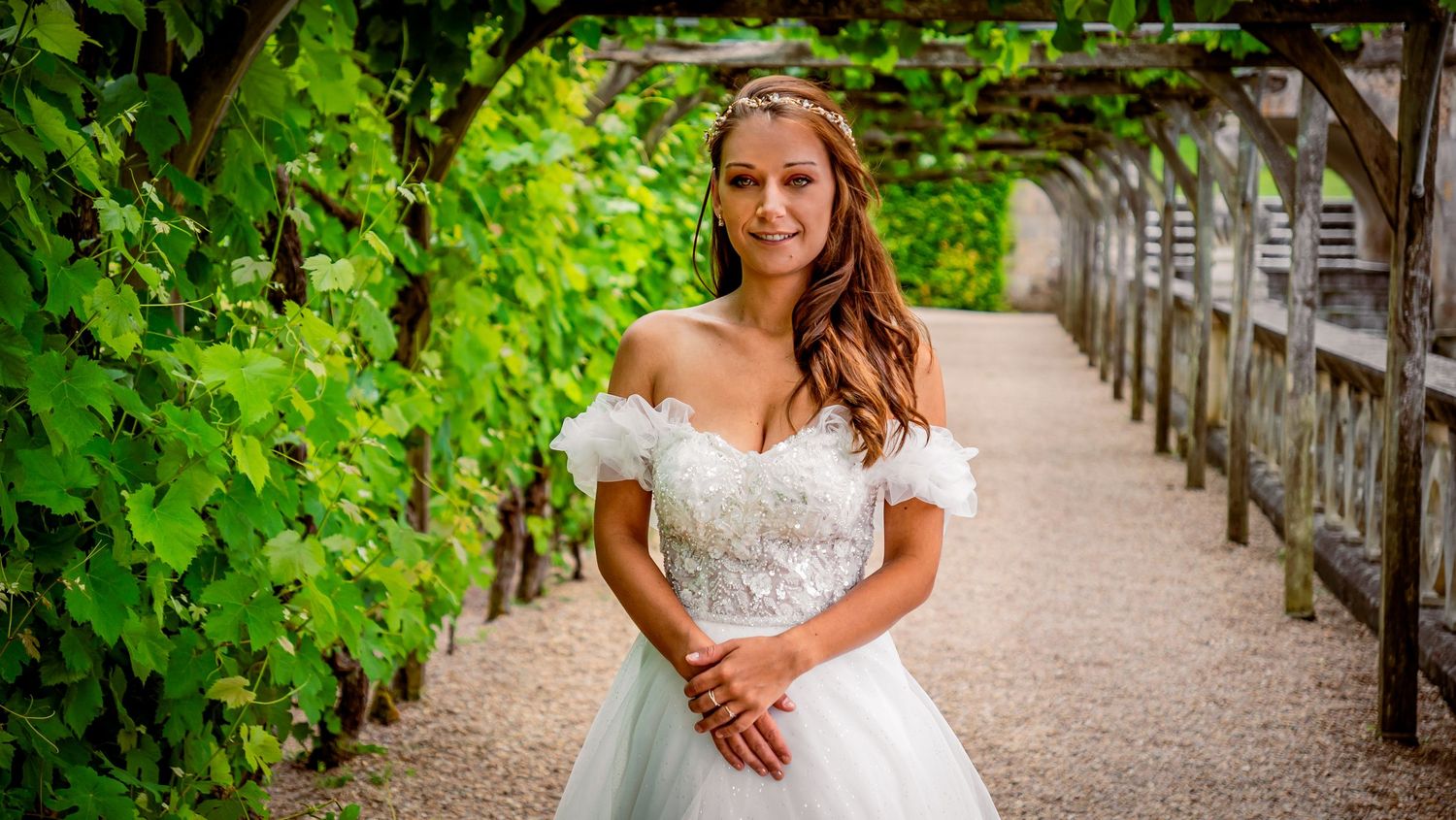 A bride in an off-shoulder white gown stands on a pathway with green vines growing along a wooden pergola.