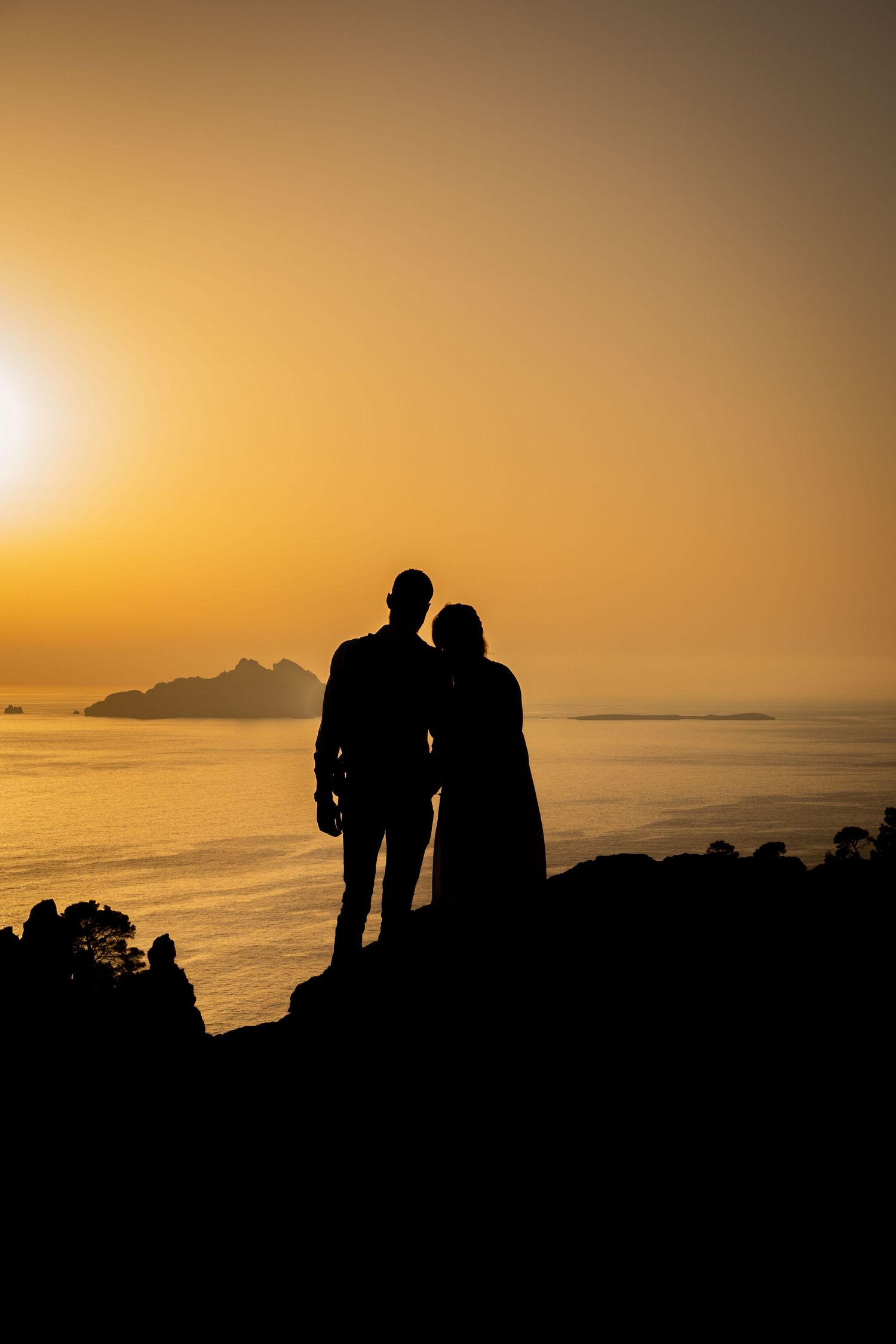 Silhouetted figures stand together against a golden sunset sky over a calm ocean horizon.