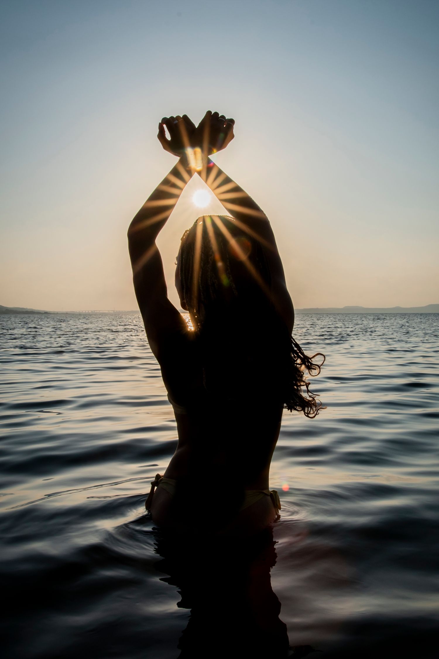 Silhouette of person in water at sunset creating diamond shape with hands above head.
