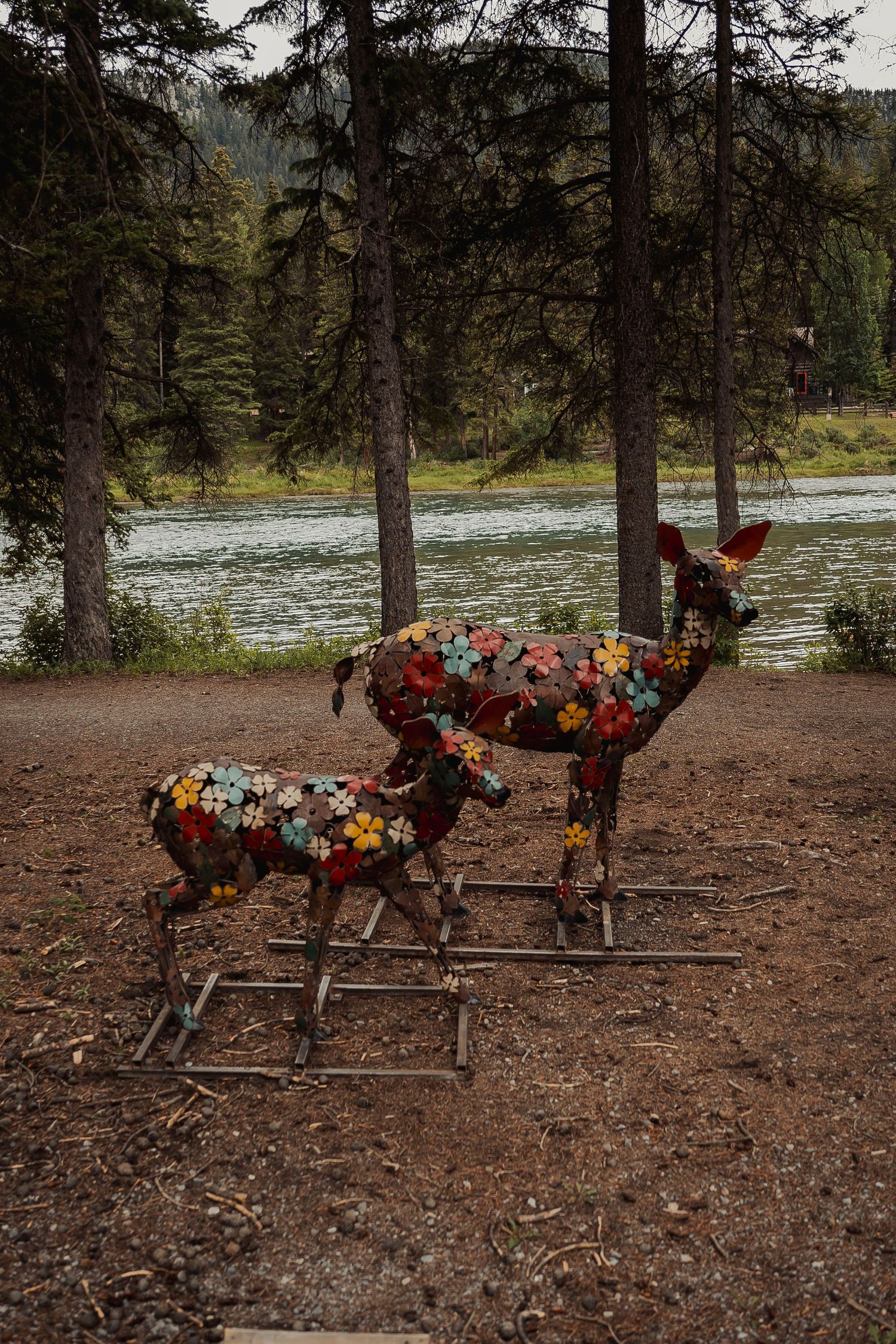 Six flower-adorned wood carvings of ducks or geese on display along a wooded riverbank scene.