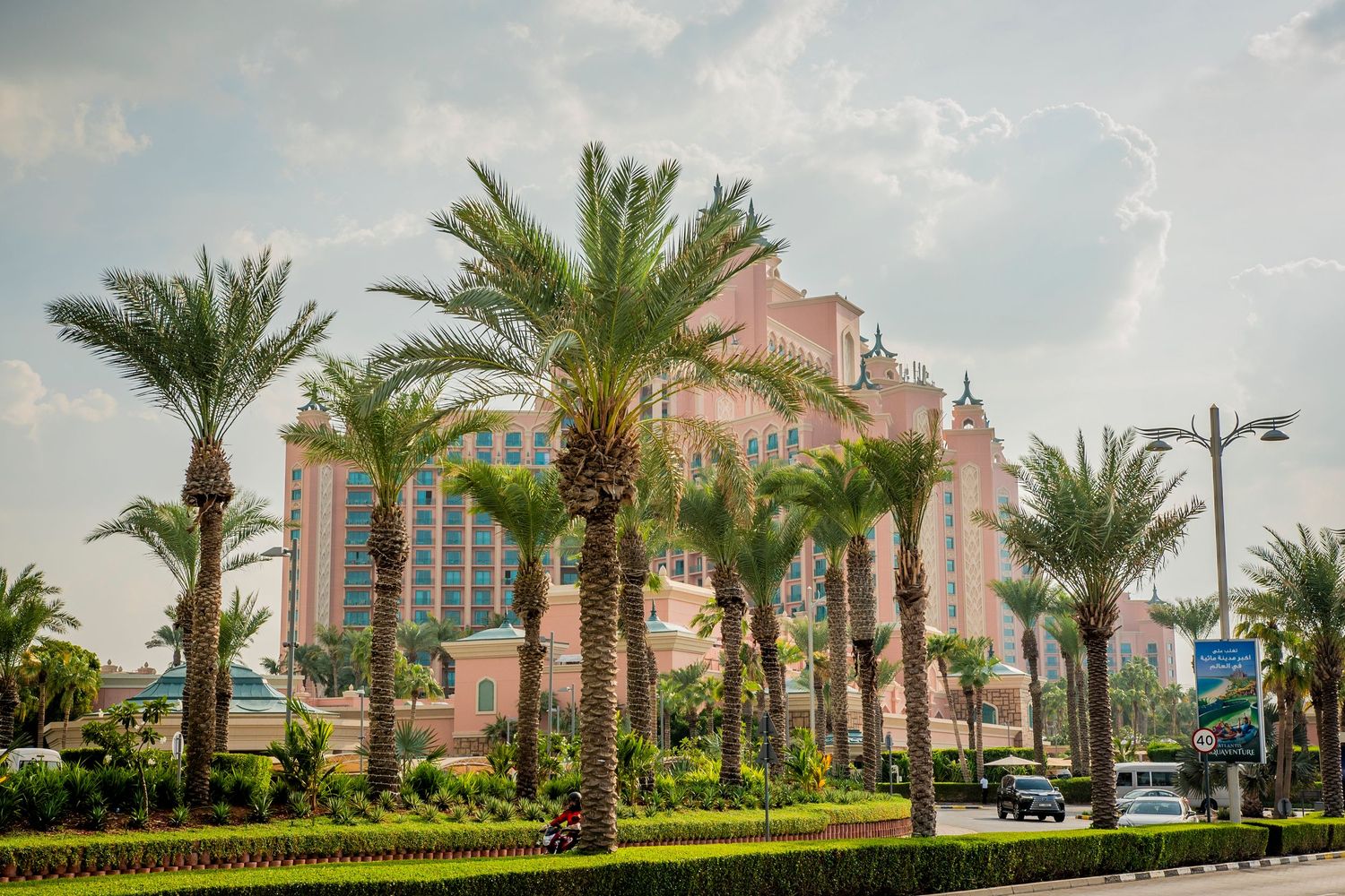 Mediterranean-style pink hotel seen through a row of palm trees against a blue sky with white clouds.