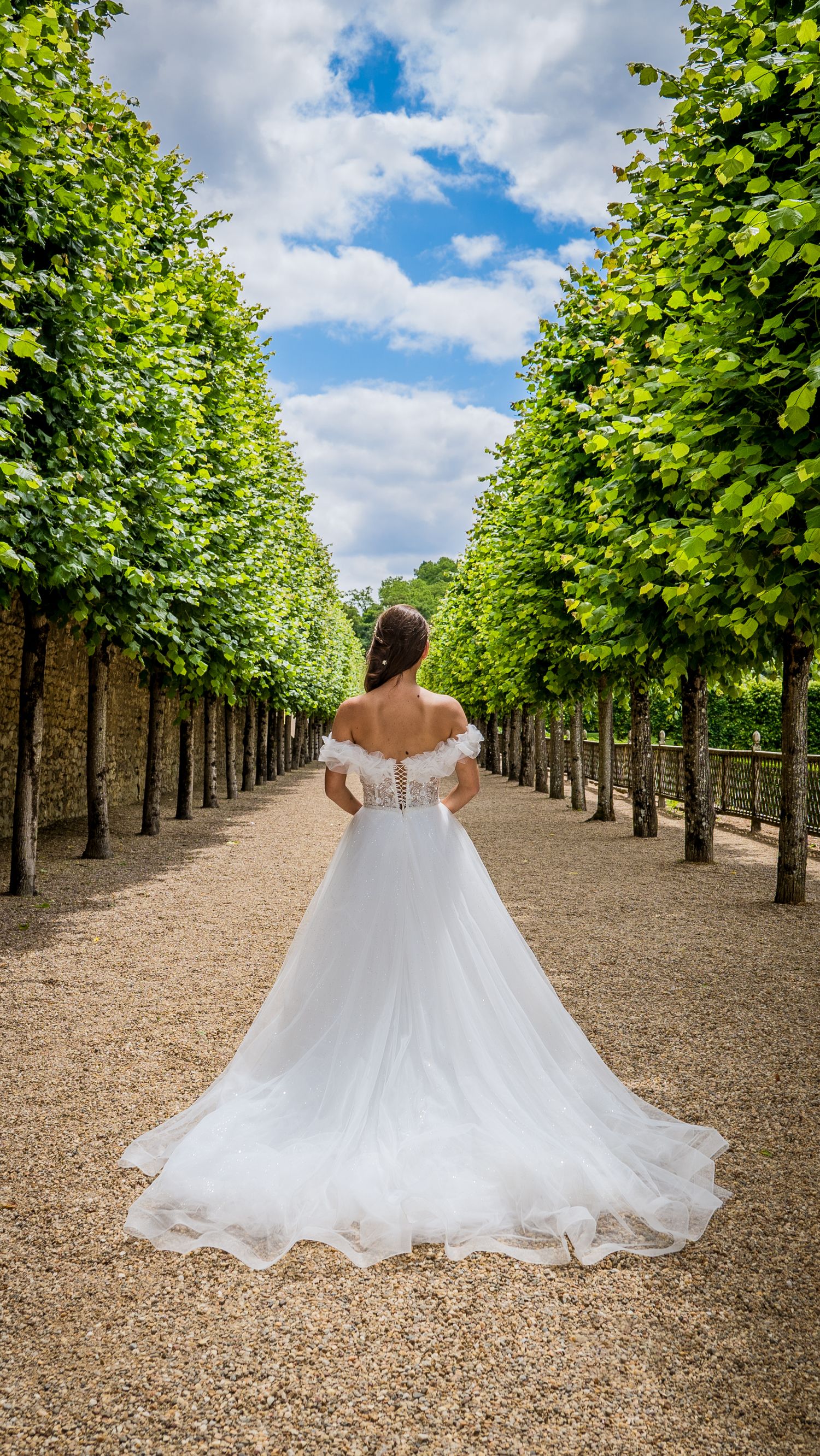 White wedding dress flowing gracefully between rows of trees in formal garden with blue sky overhead.
