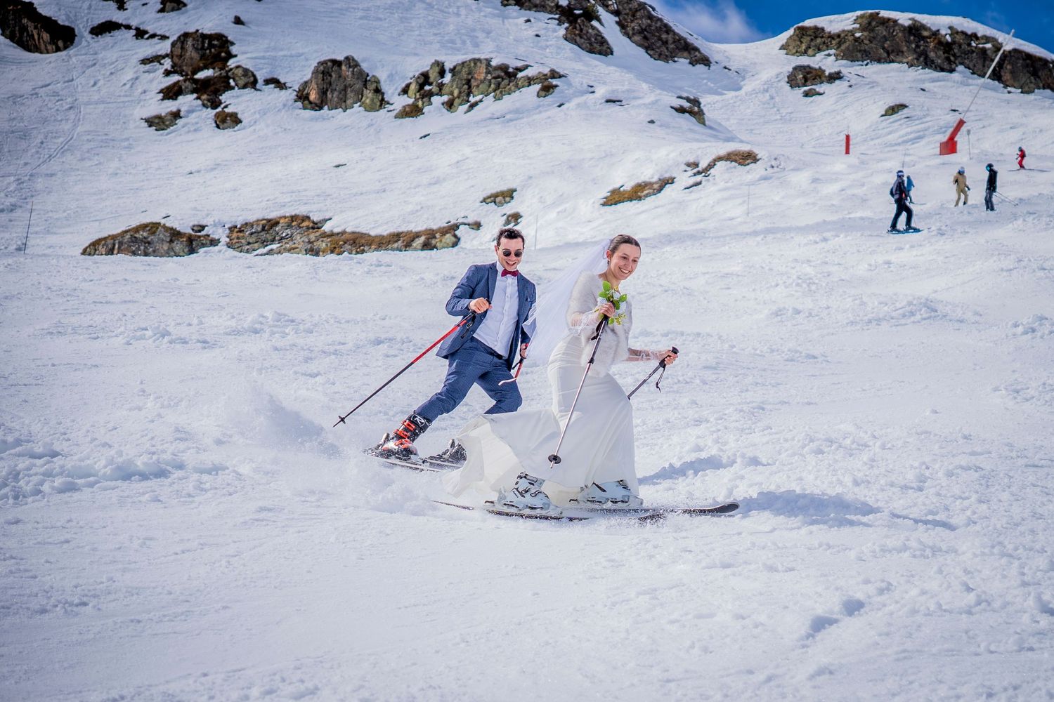 Two skiers gliding down a snowy mountain slope with rocky peaks and blue sky visible in the background.