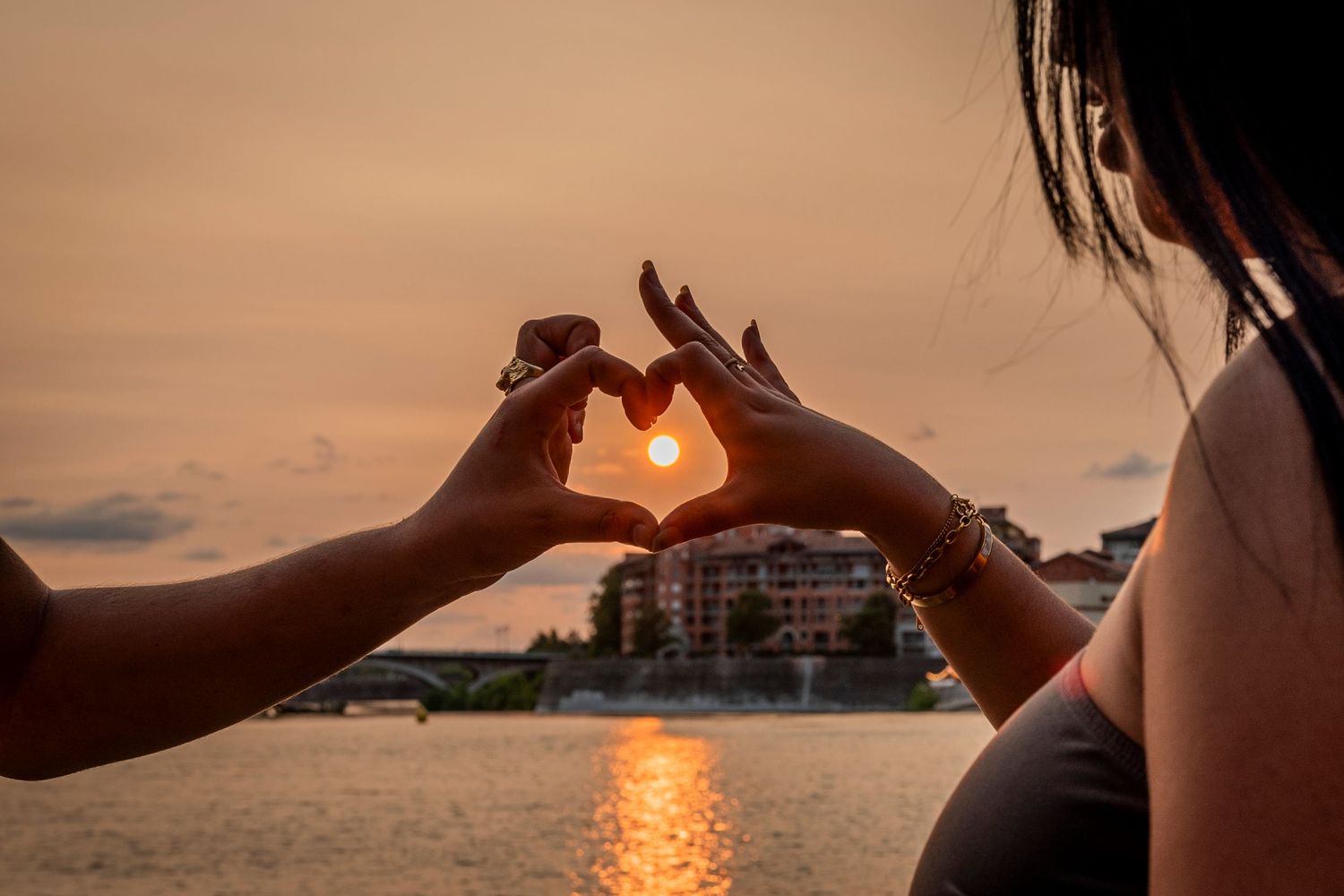 Two hands form a heart shape against a sunset over water with the sun glowing through the silhouette.