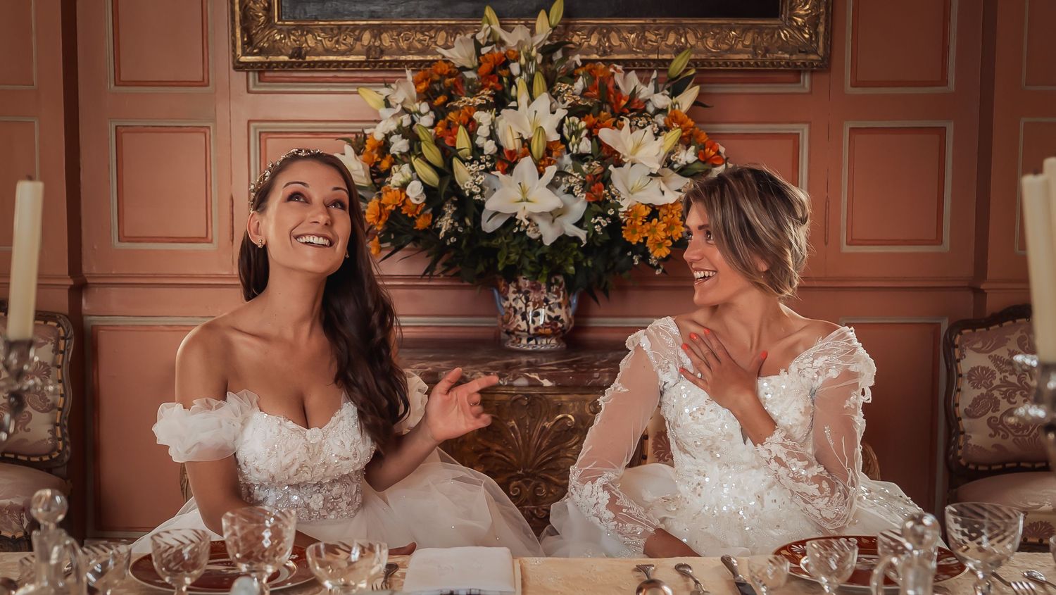 Two people in white lace gowns share a joyful moment while seated at an elegantly decorated dining table.
