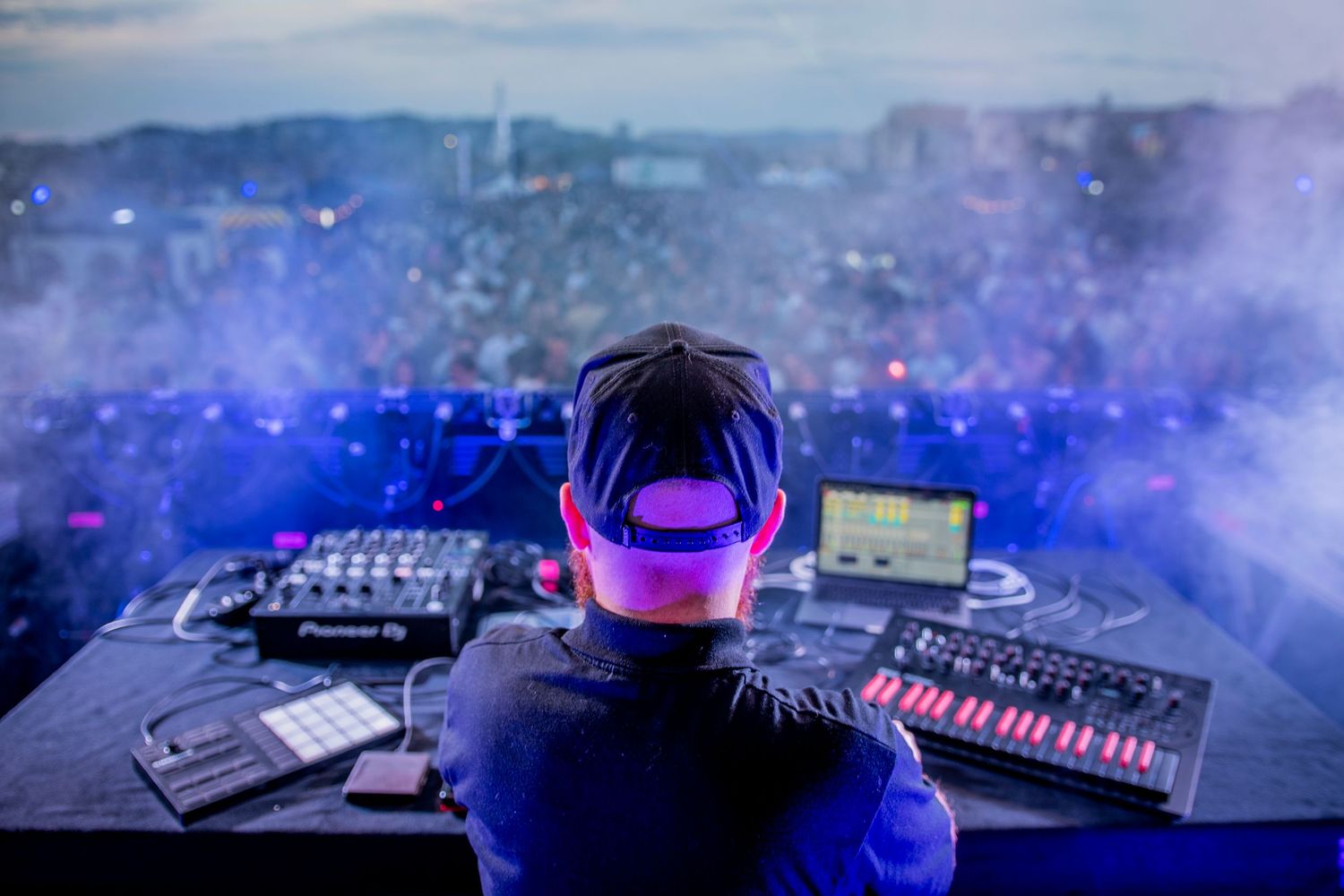 A DJ performs at a music festival with glowing equipment and purple lights while a crowd fills the smoky background.