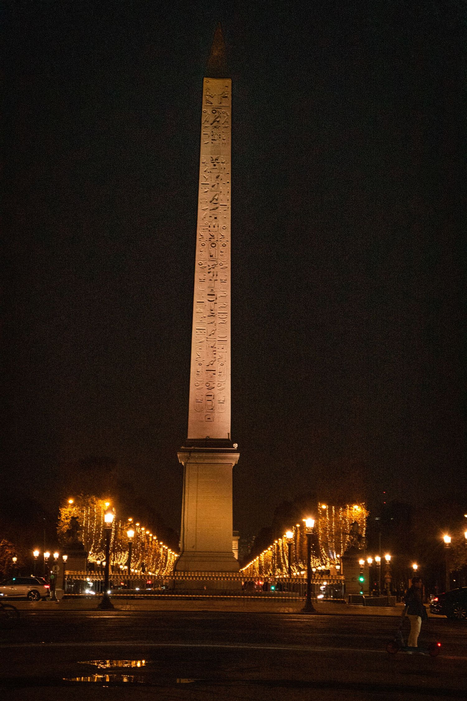 Illuminated obelisk monument stands tall at night surrounded by glowing street lamps along a grand avenue.