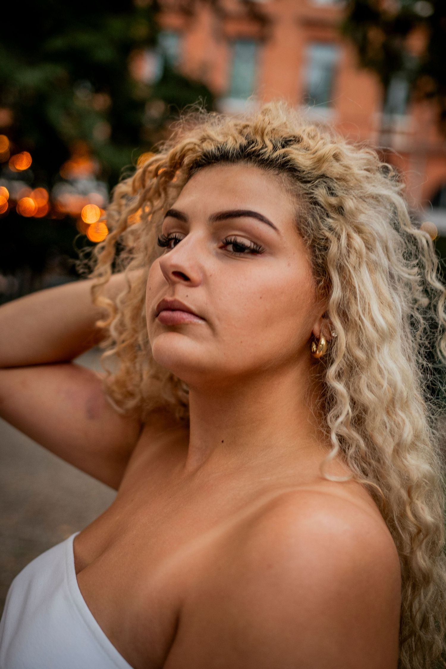 Portrait of someone with voluminous blonde curly hair and gold earrings in golden hour lighting against a brick wall.