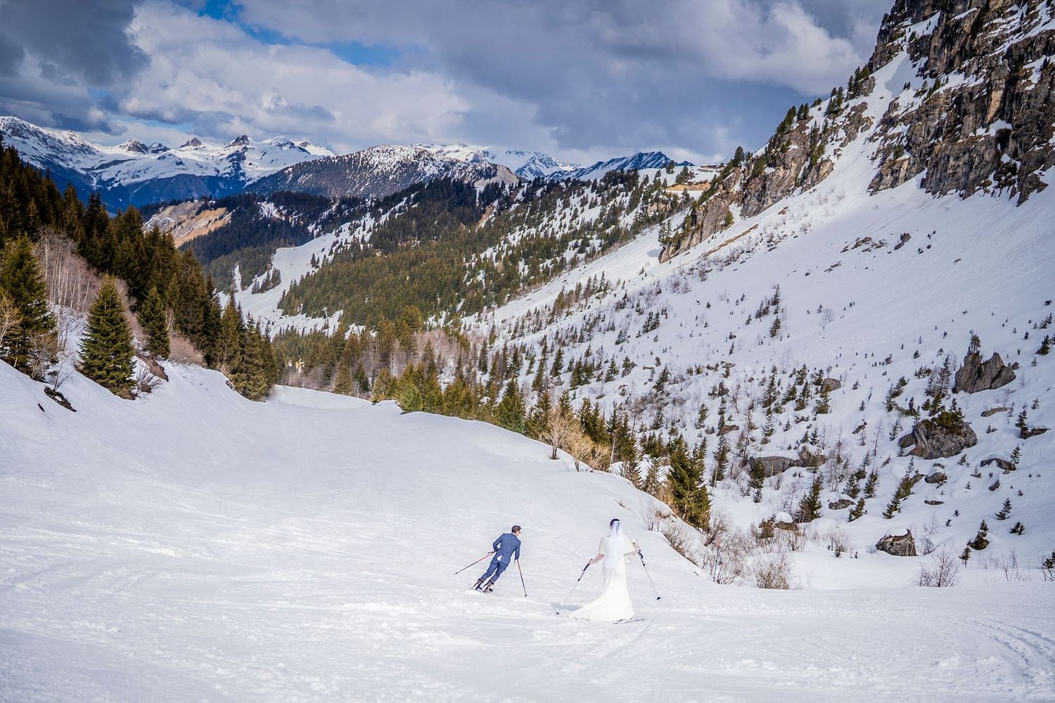 A skier descends a snowy slope surrounded by majestic mountain peaks and evergreen trees in a winter landscape.