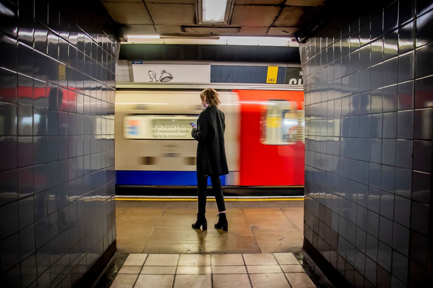 Person walks past dark tiled walls in an underground subway station with illuminated train in background.