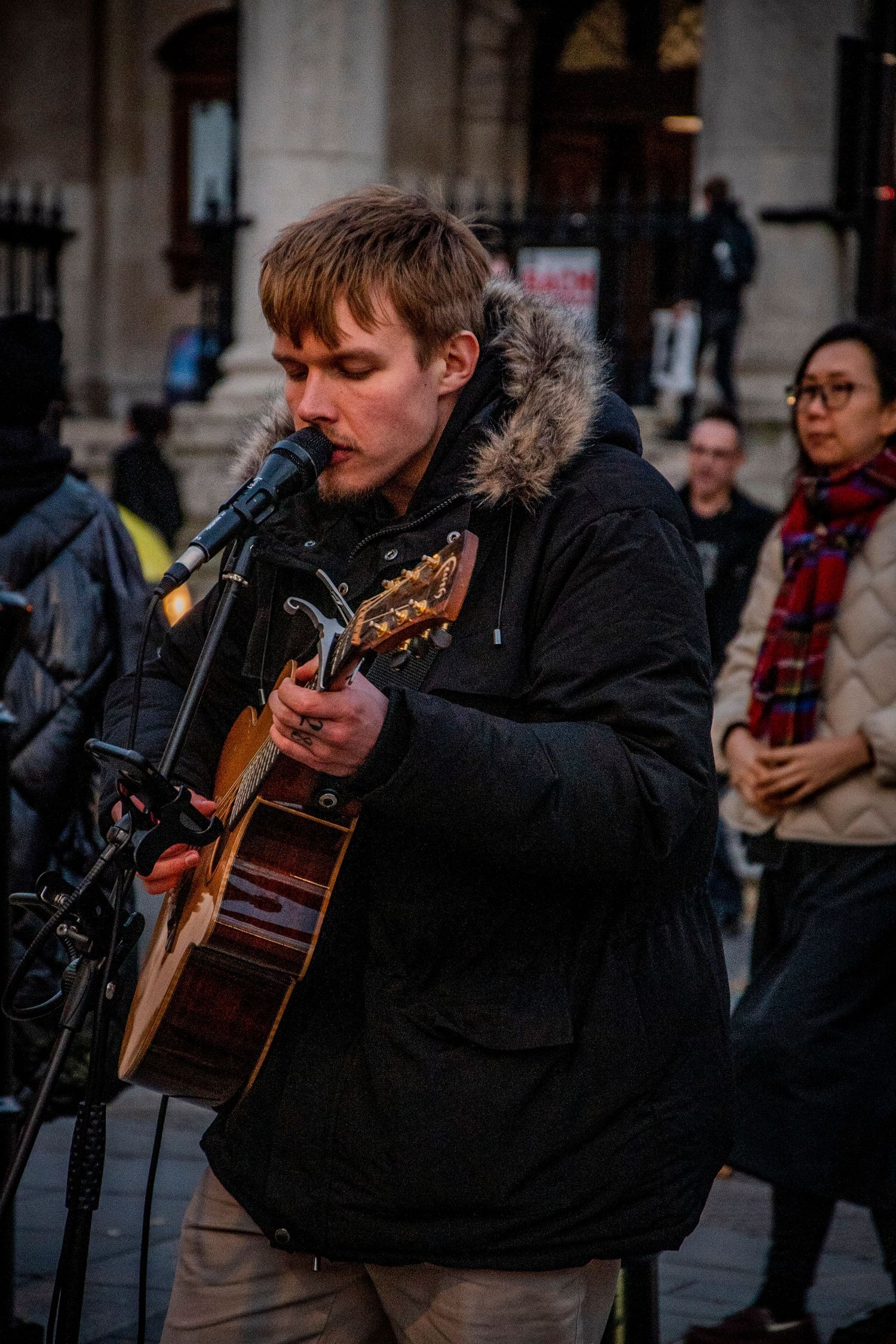 A street musician performs with a guitar outside in cold weather wearing a winter coat with fur-lined hood.