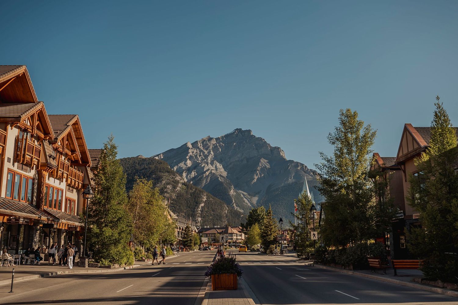 Row of mountain lodge buildings with large windows and wooden beams nestled beneath towering rocky peaks.