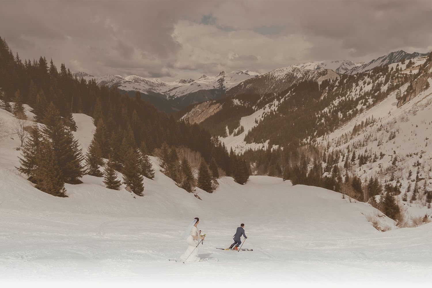 Two skiers descending a snowy slope in a majestic mountain valley with evergreen forest and snow-capped peaks.