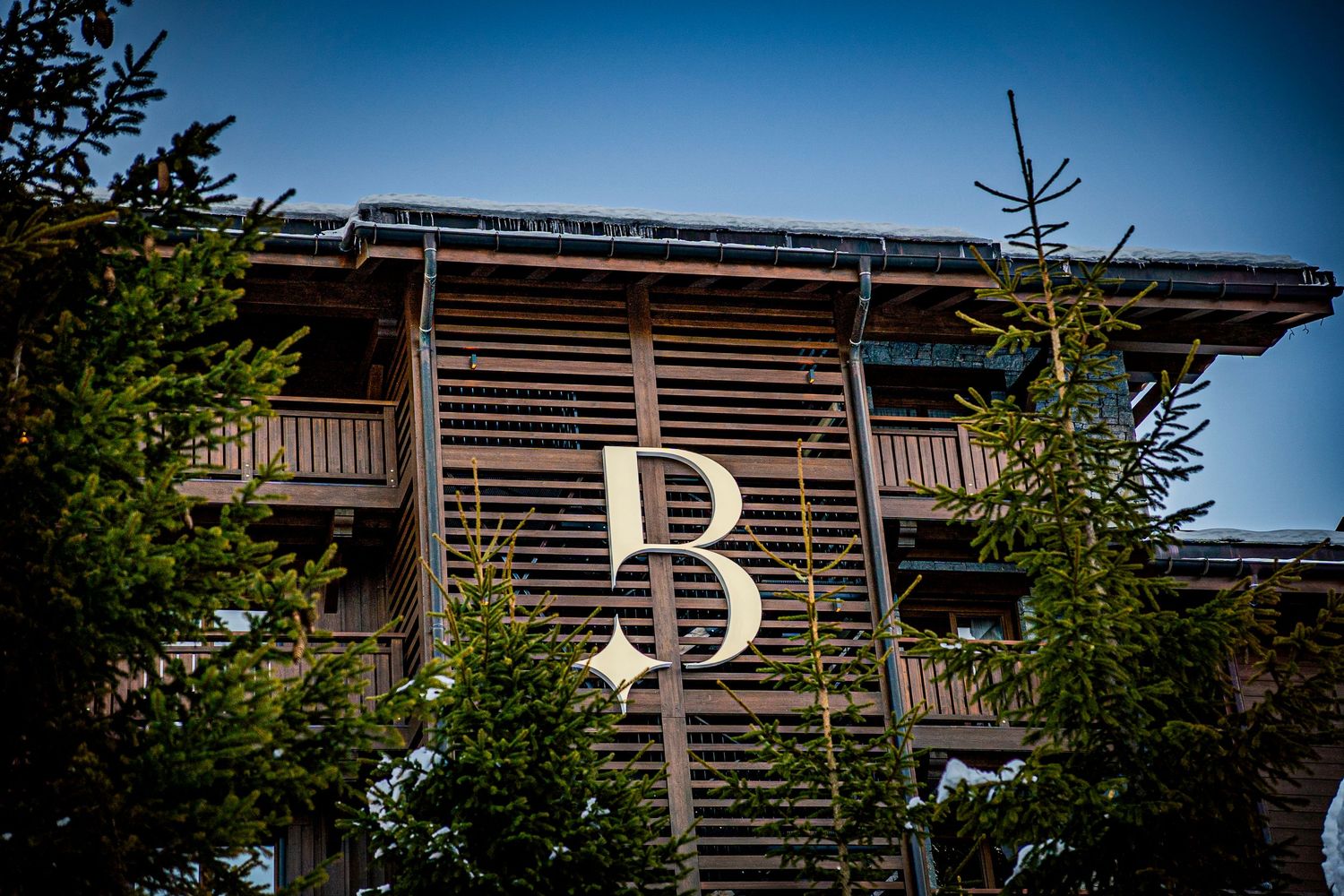 Modern wooden apartment building exterior with slanted panels and balconies surrounded by evergreen trees against blue sky.