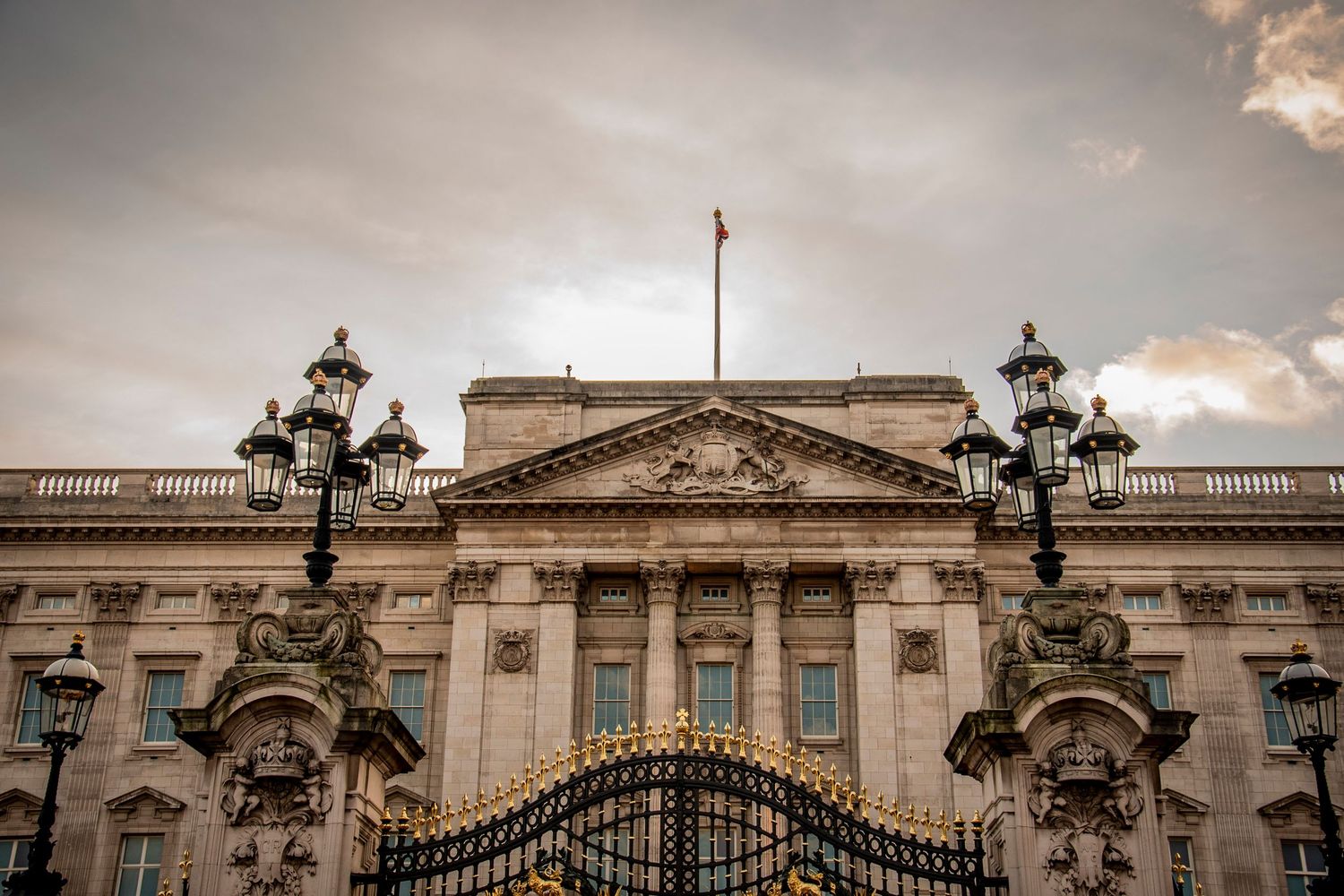 Ornate architectural details of Buckingham Palace gates and facade with dramatic clouds in the background.