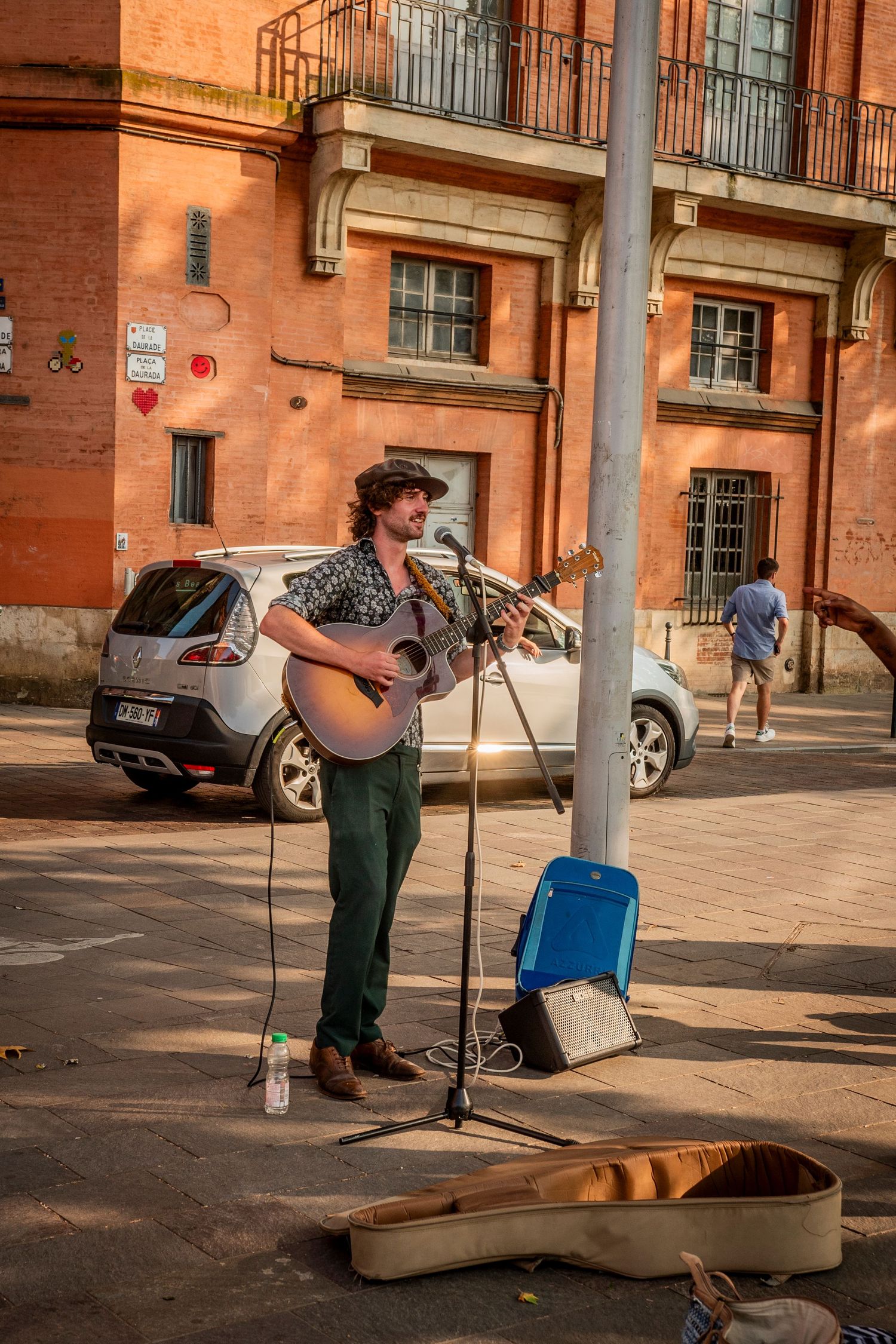 Street musician performs on guitar with open case for donations on a sunny sidewalk near a brick building.