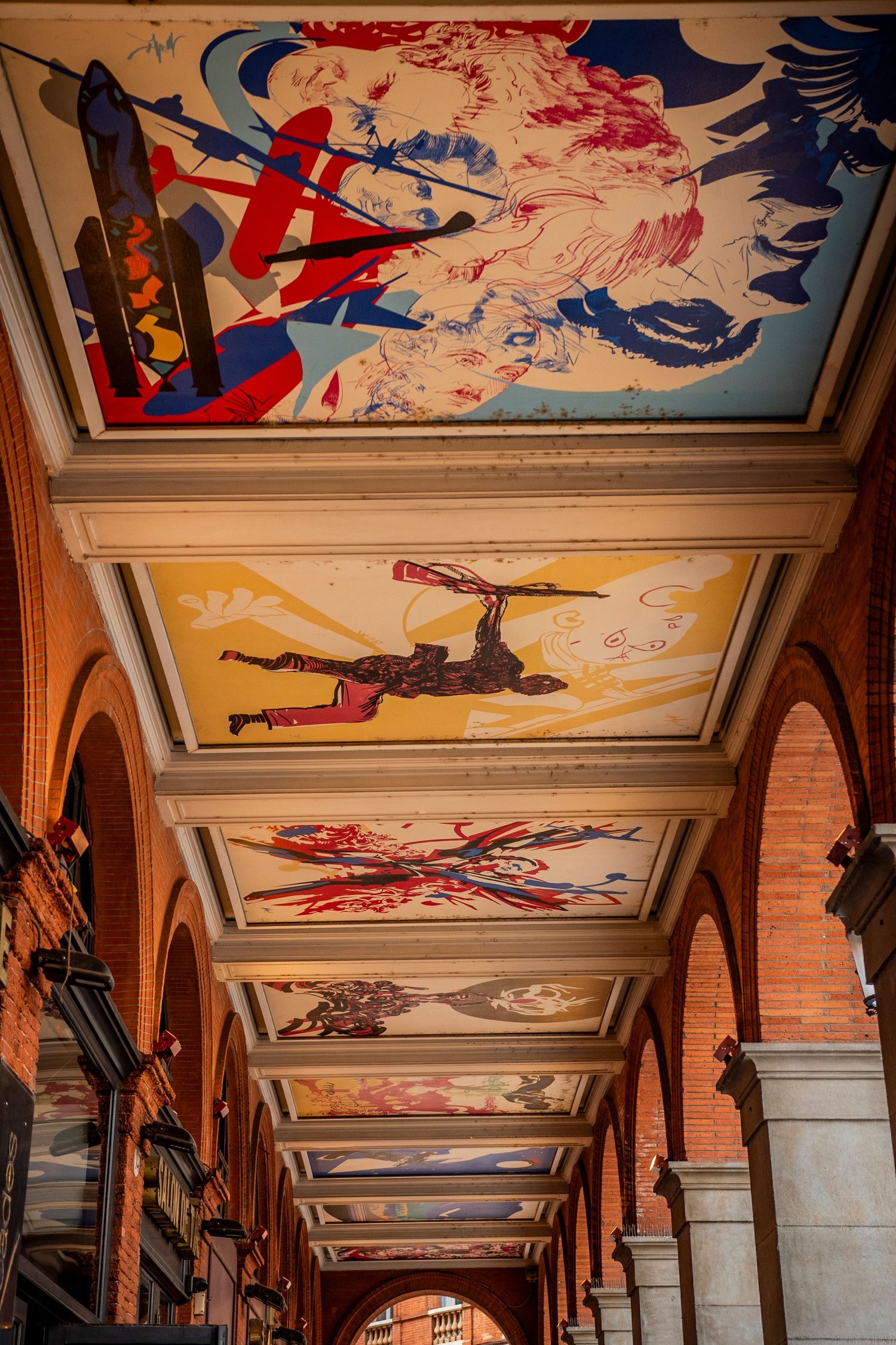 Colorful ceiling murals depicting planes and aircraft in a historic arcade hallway with red brick arches and columns.