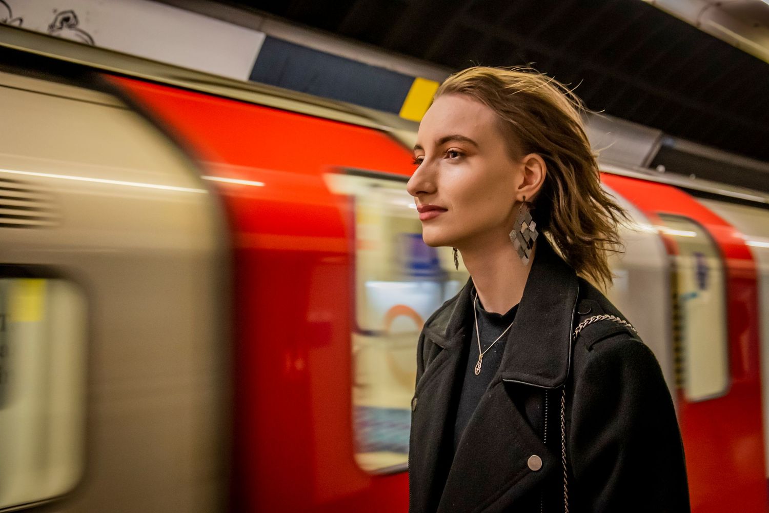 Figure in black coat stands contemplatively on subway platform as red train car blurs past in motion.