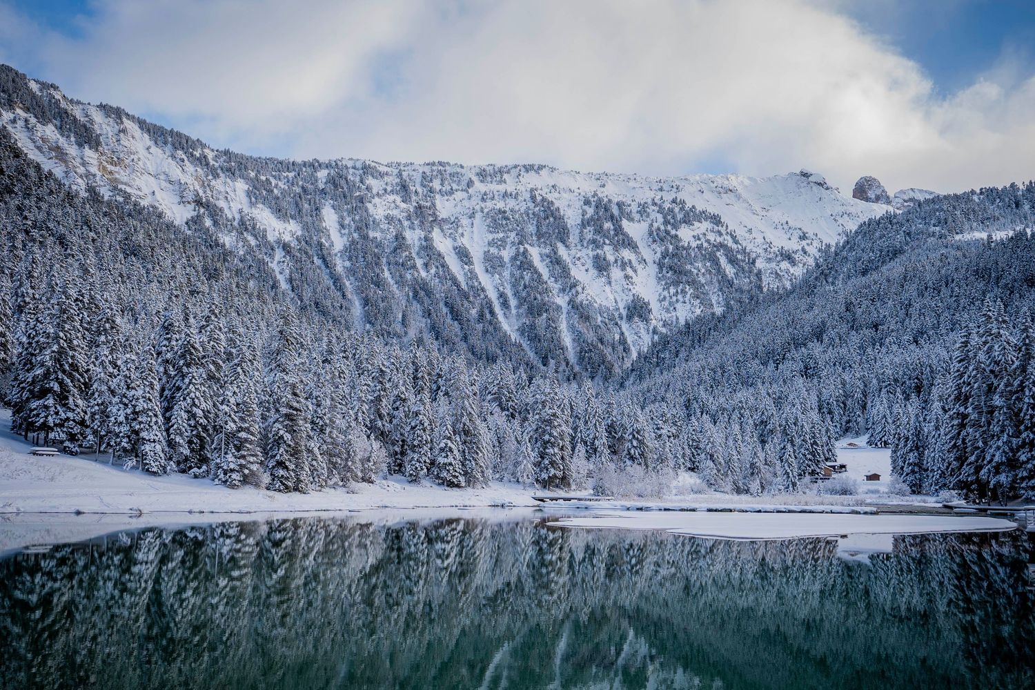 Snow-covered mountains and evergreen forests reflect in a pristine alpine lake under cloudy winter skies.