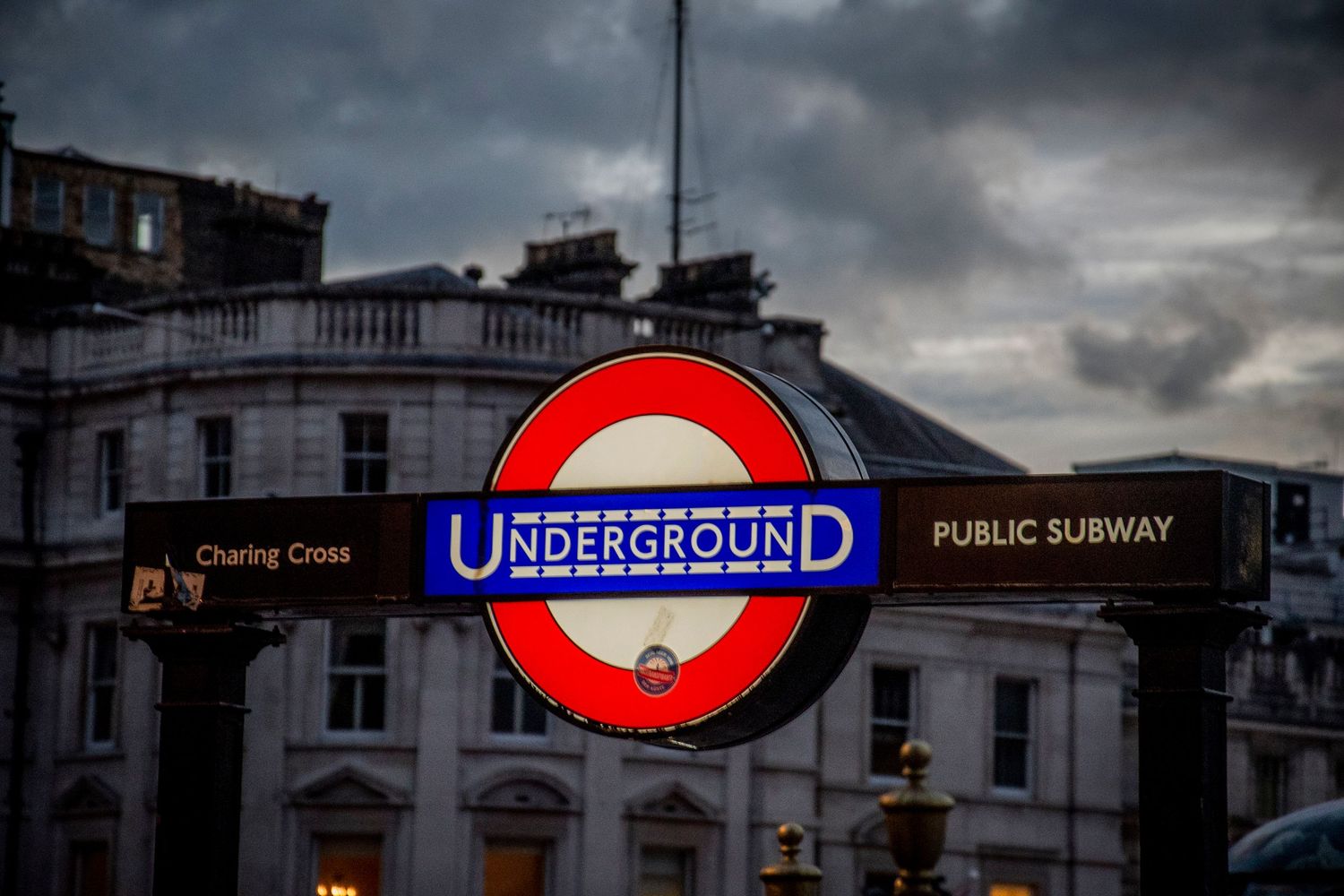 Iconic London Underground sign glows against moody sky with historic buildings in the background.