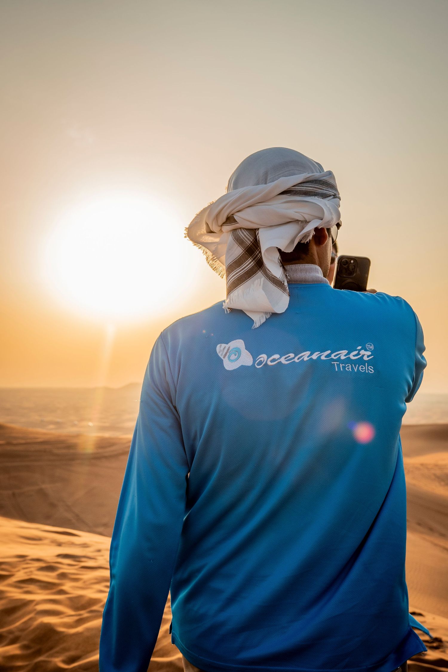 Person wearing traditional Middle Eastern headwear and tourism company shirt gazes at desert sunset landscape during safari.