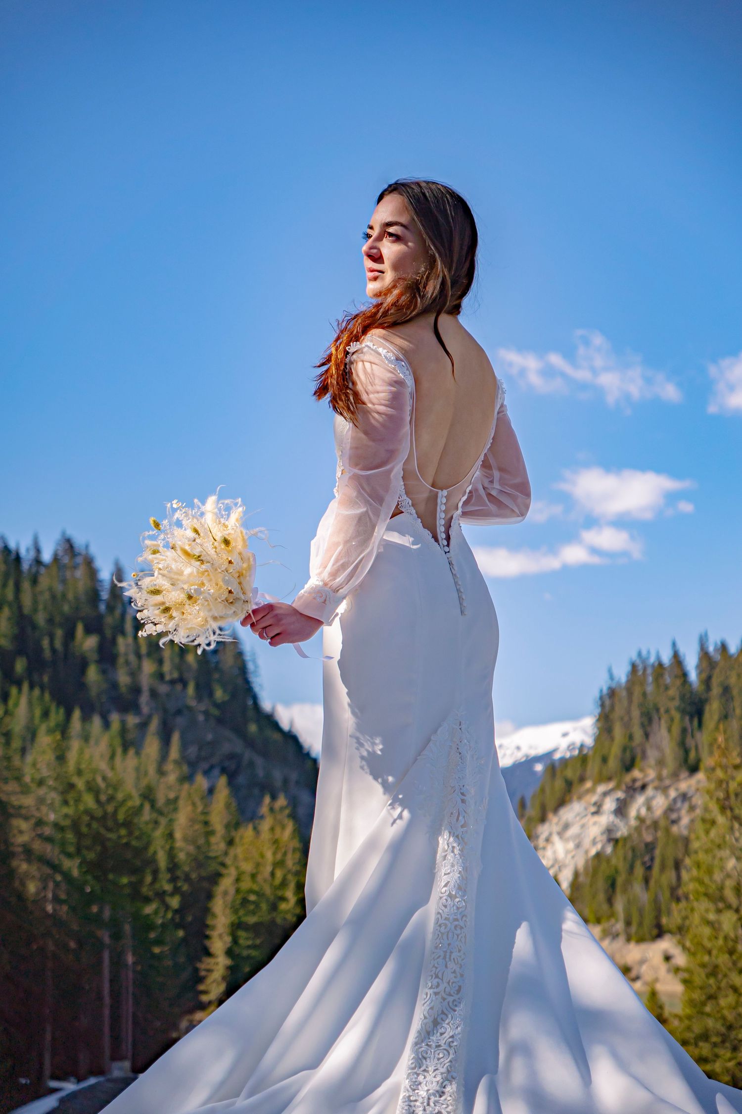 Bride in elegant white long-sleeve dress holding dried bouquet against mountain landscape with evergreen trees and blue sky.