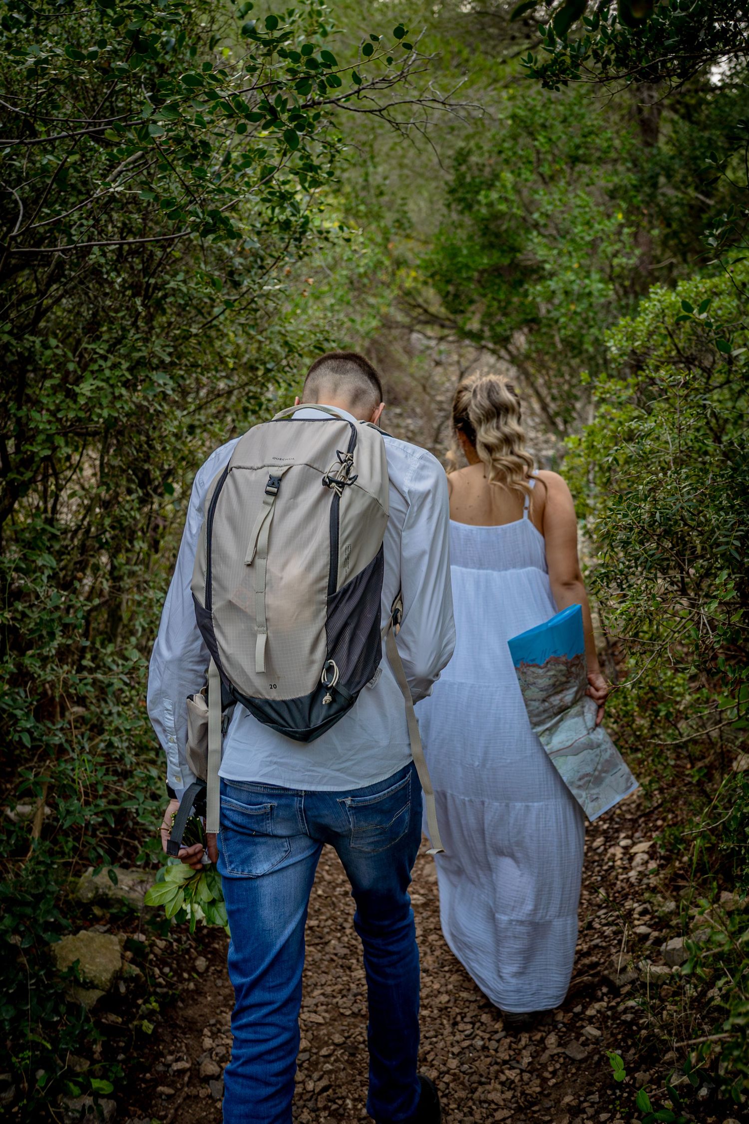 Two hikers exploring a shaded forest trail together, one carrying a backpack and another wearing a flowing white dress.