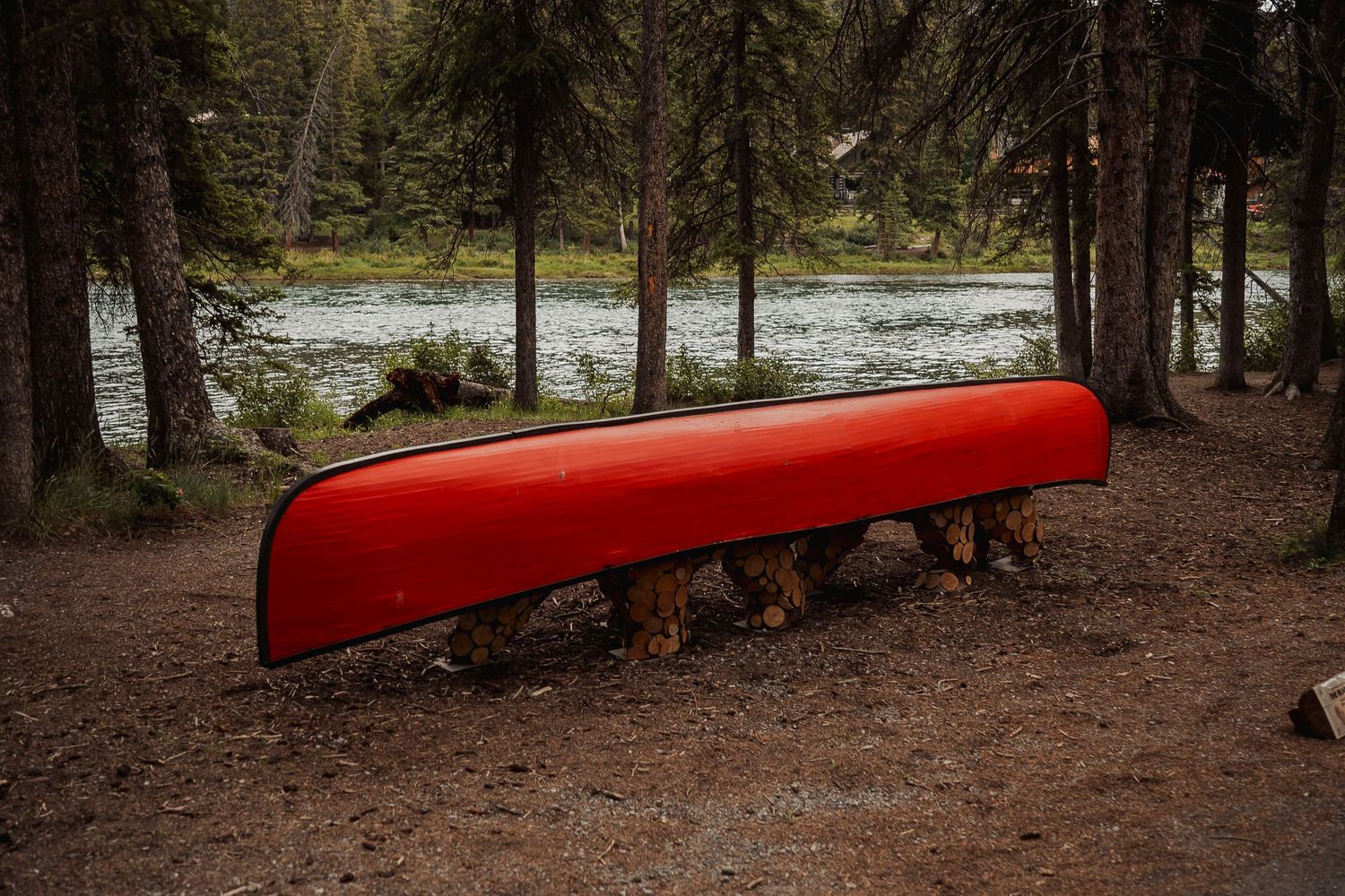 A bright red canoe rests upside down on logs in a wooded forest area near a serene lake.