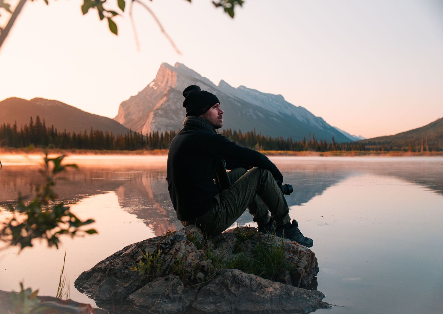 Person sitting on rocky overlook watching sunset over tranquil mountain lake with reflection in calm waters.