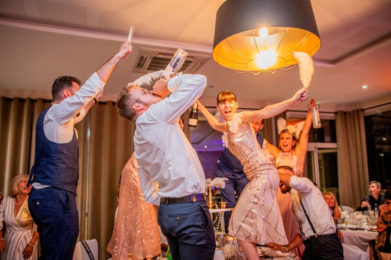 Wedding guests enthusiastically dancing together with raised arms under a warm pendant light at an indoor reception party.
